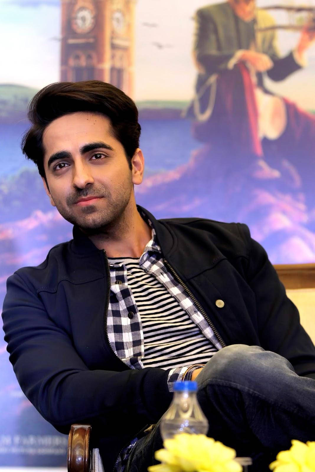 ayushmann khurrana 桌布 壁紙 — 一名身穿黑色外套和條紋襯衫的男子，隨意地坐在電影海報背景前。