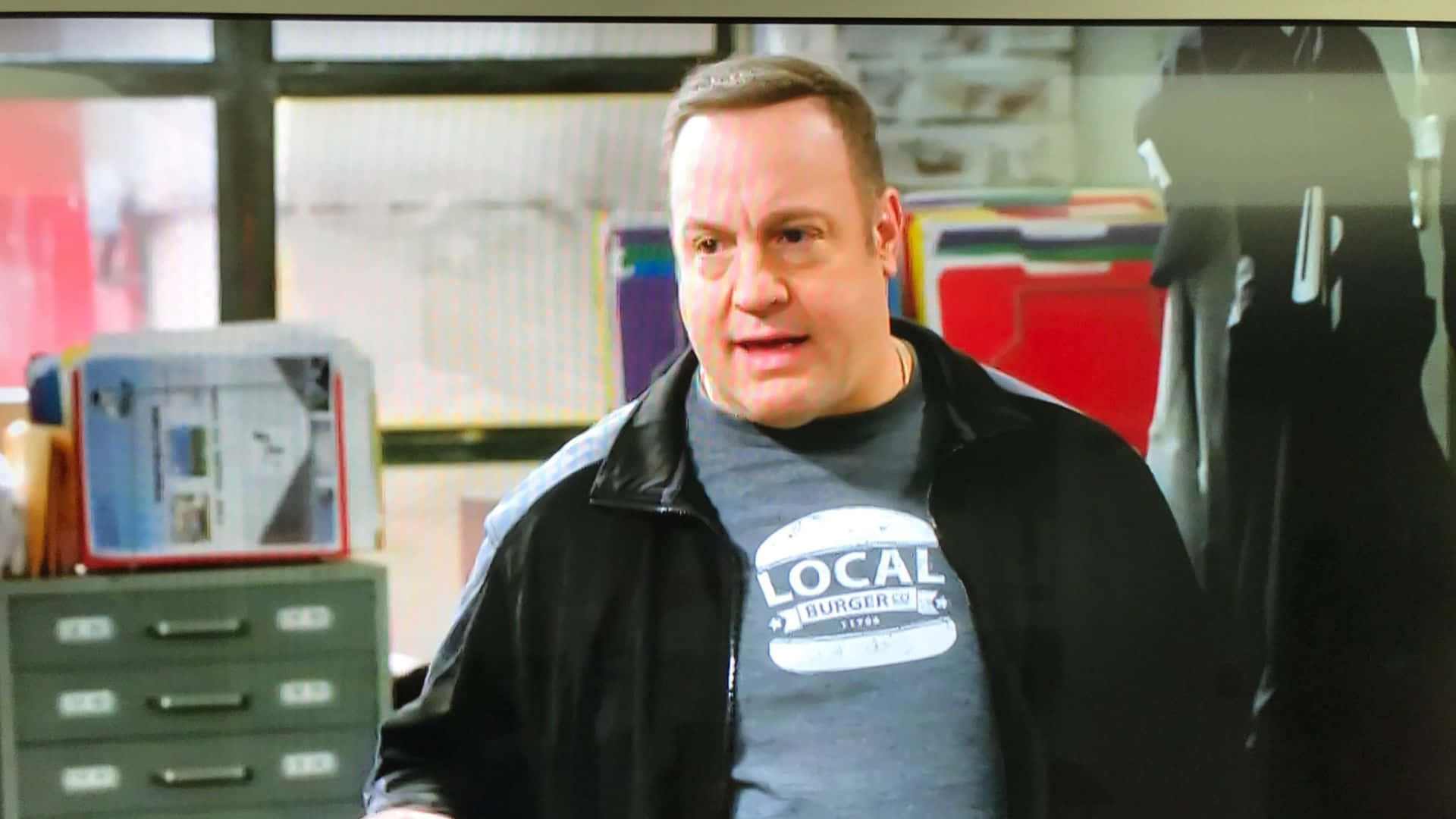 Kevin James 桌布 壁紙 — 一名身穿黑色外套和灰色「Local Burger Co.」T恤的男子站在雜亂的辦公室裡，身後有文件櫃和彩色文件夾。
