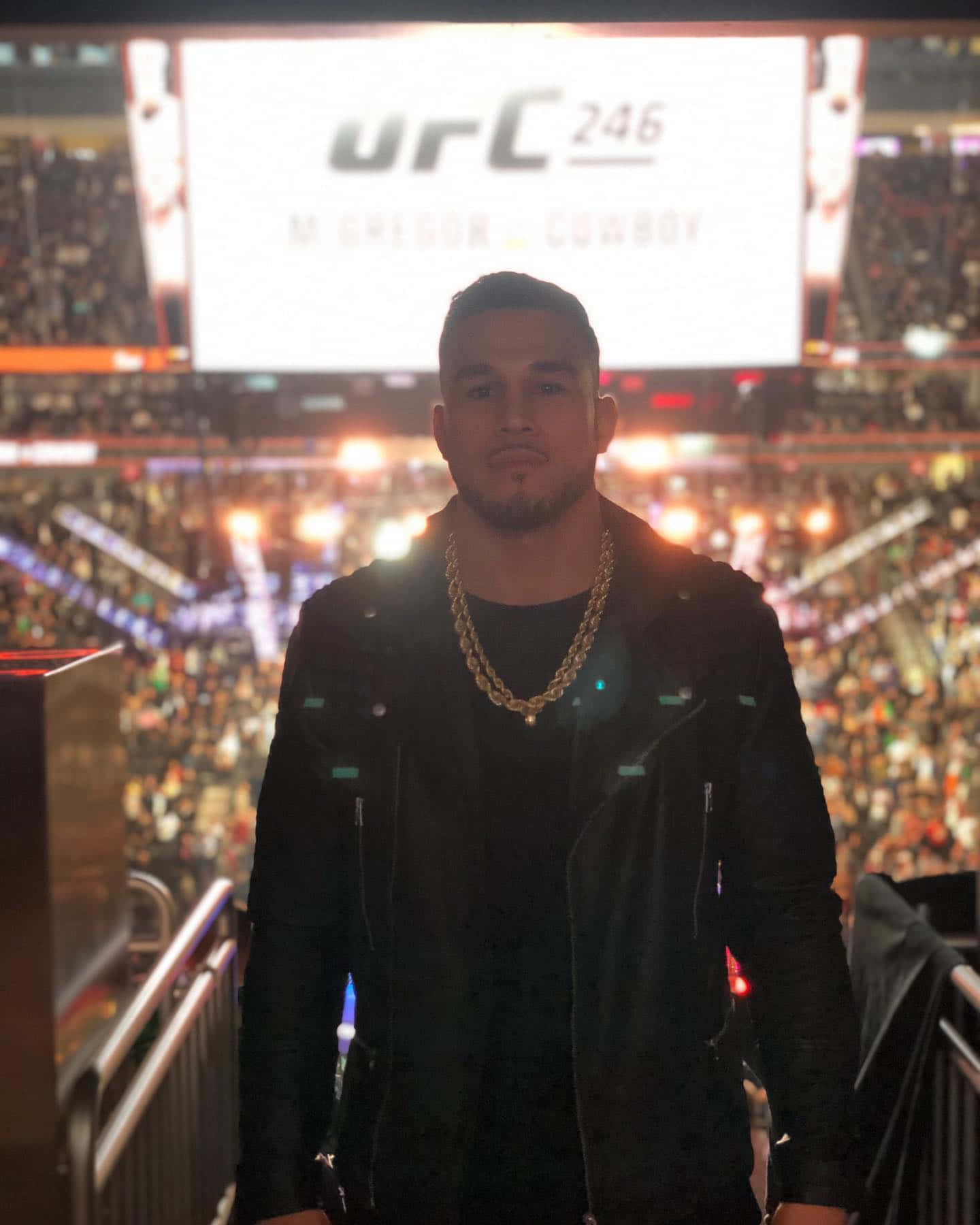 MMA 桌布 壁紙 — 一名身穿黑色外套和金色鏈條的男子自信地站在擁擠的競技場中，身後有一個大屏幕顯示 'urC 246 M CHIGOR COWBOY'。