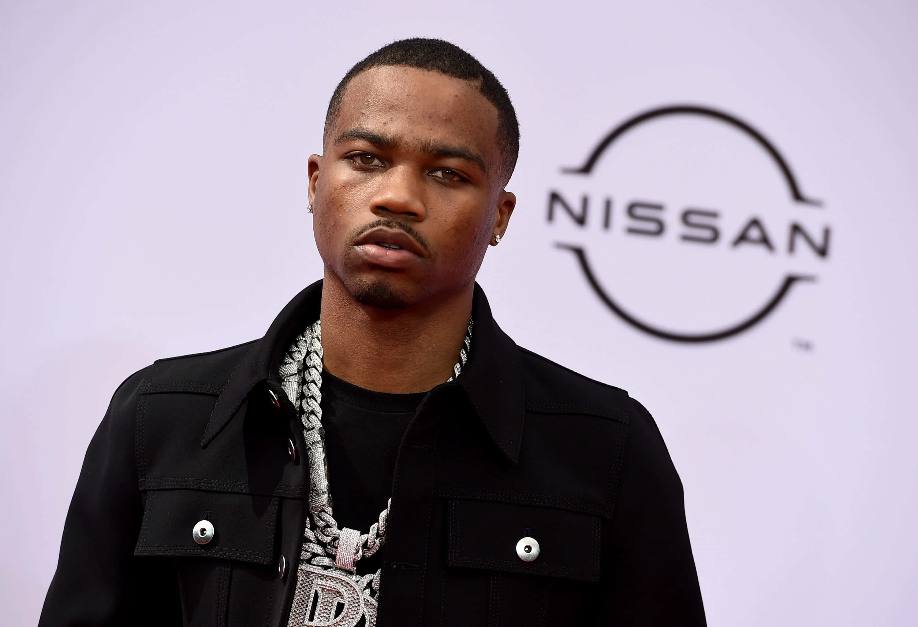 Roddy Ricch 桌布 壁紙 — 一名身穿黑色外套和銀鏈的男子自信地站在日產標誌背景前。