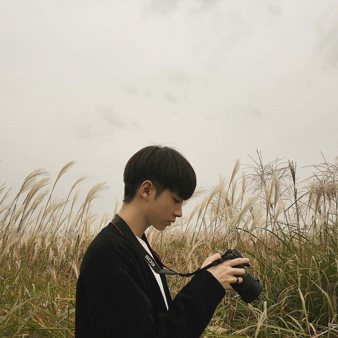 Holding Camera 桌布 壁紙 — 一名身穿黑色外套的年輕男子站在高高的金色草叢中，手持相機，天空多雲。