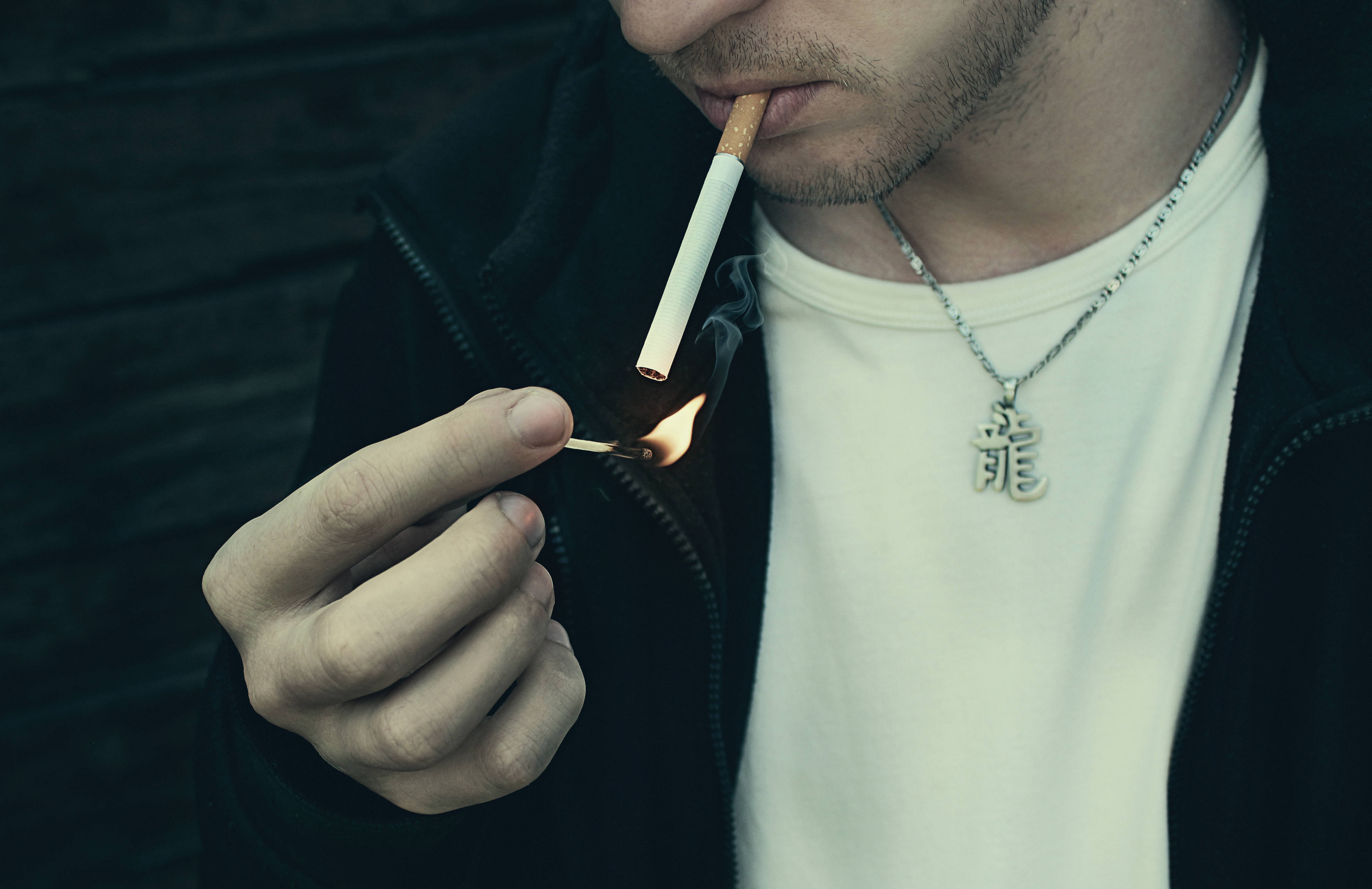 Smoking 桌布 壁紙 — 一名身穿黑色外套的男子，在深色磚牆前用火柴點燃香菸。