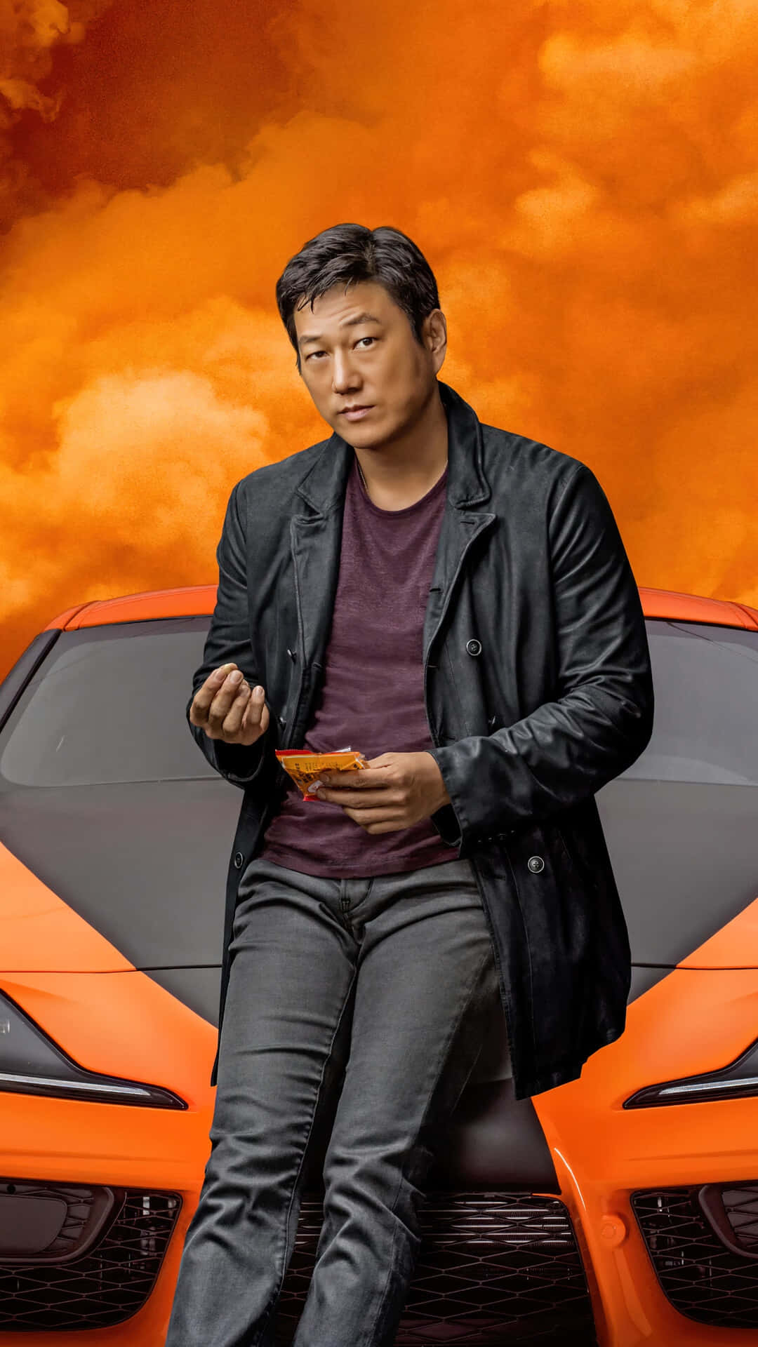 Fast And Furious 9 桌布 壁紙 — 一名身穿黑色外套的男子，在火紅的橙色天空下，隨意地坐在一輛充滿活力的橙色跑車的引擎蓋上。