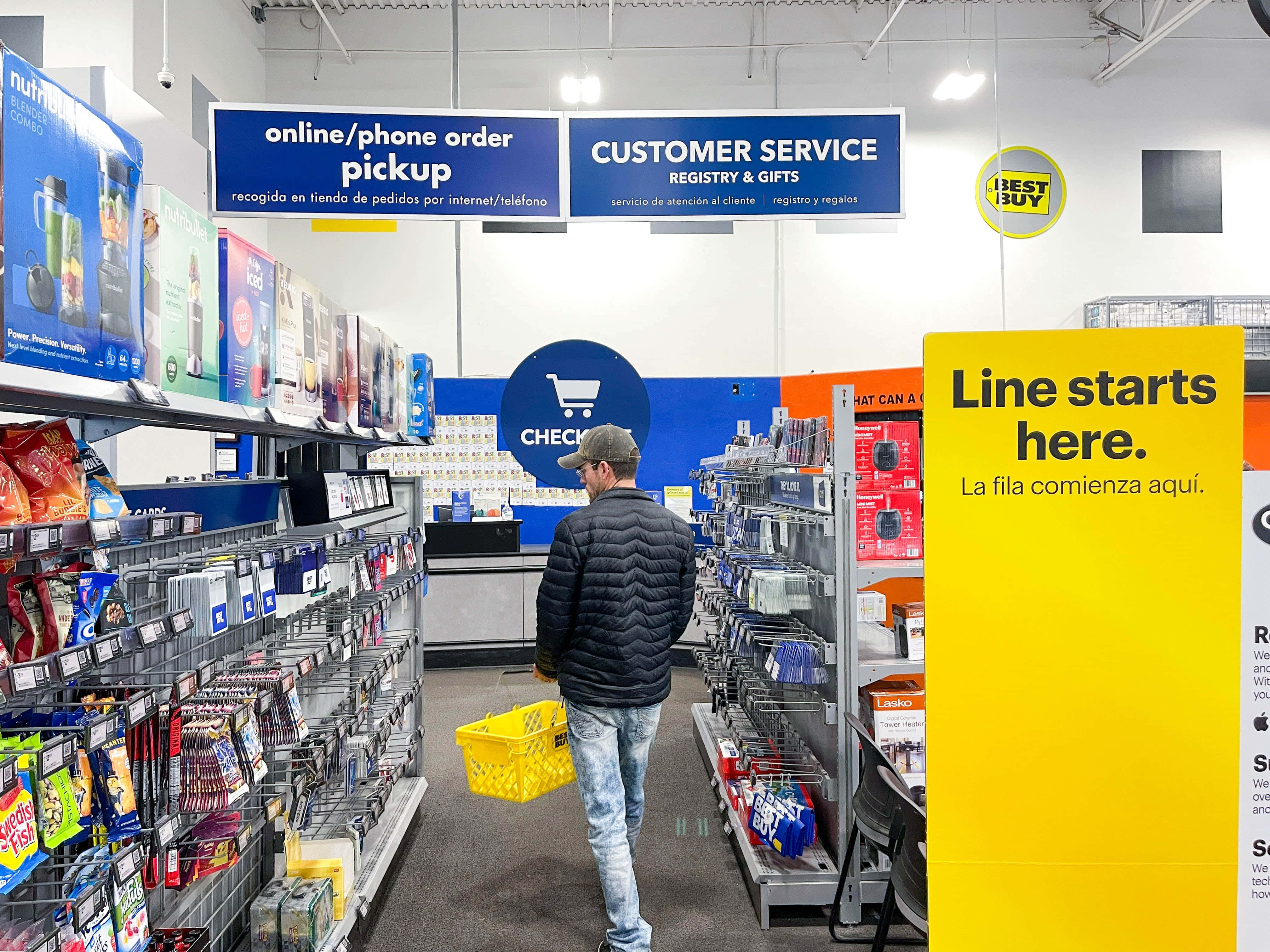 Best Buy 桌布 壁紙 — 一名身穿黑色外套的男子在Best Buy的通道中拿著黃色購物籃走向收銀台，頭頂有明亮的標牌。