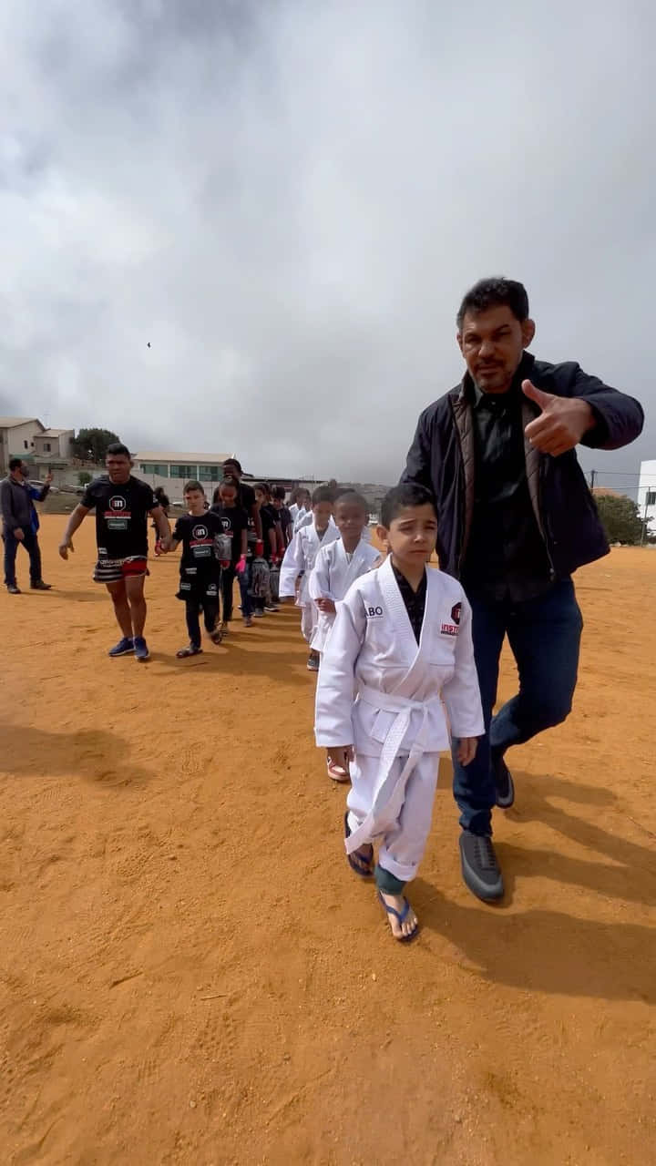 小孩動圖 桌布 壁紙 — 一名身穿黑色外套的男子在陰天下的塵土飛揚場地上，與一名身穿白色武術道服的小男孩同行時豎起大拇指。