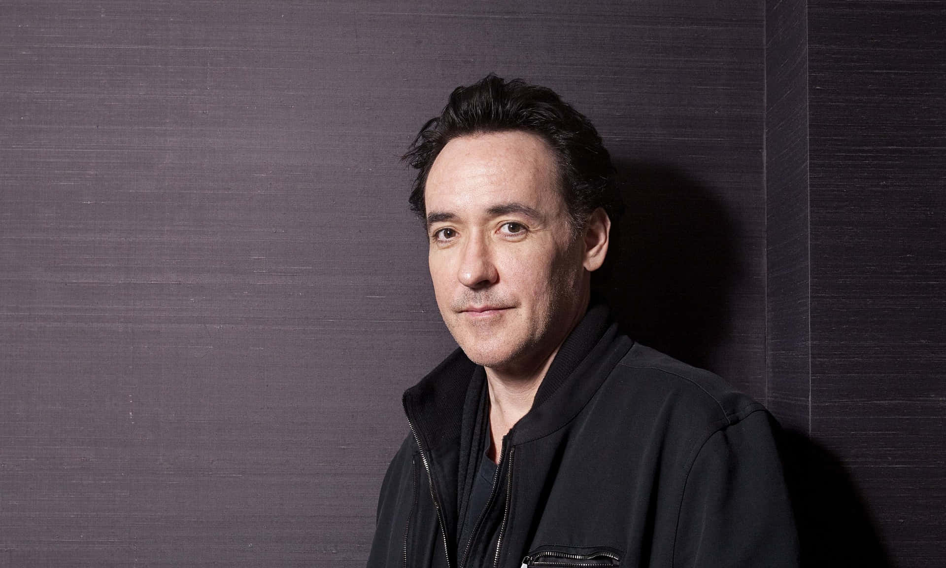 John Cusack 桌布 壁紙 — 一名身穿黑色外套的男子靠在一面深色紋理牆上，柔和的光線凸顯他的臉龐。