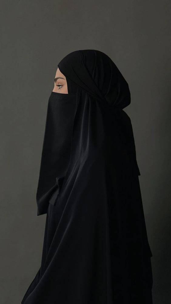 muslim 桌布 壁紙 — 一名身穿黑色尼卡布的女性側面站立，背景為柔和的灰色，她的眼睛可見，目光向前。