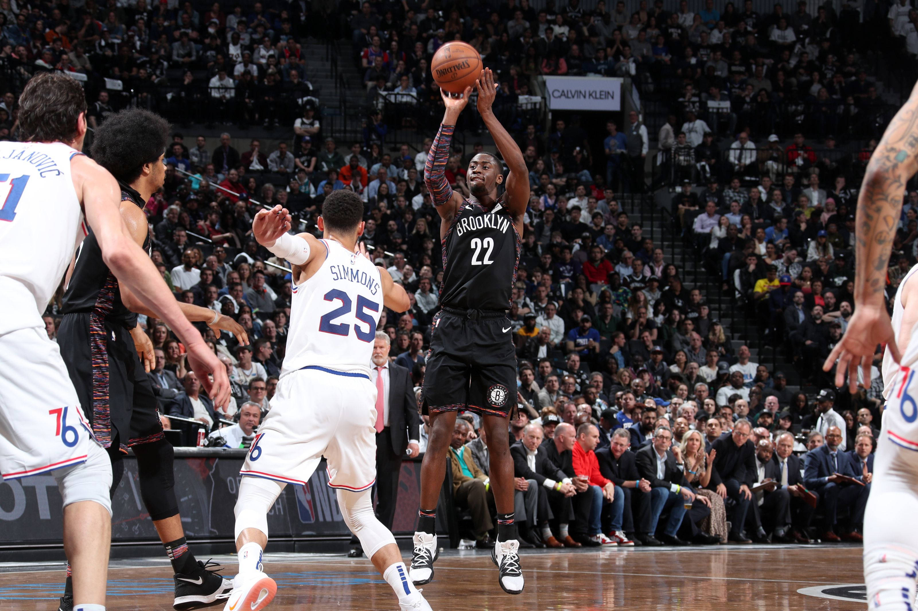 caris levert 桌布 壁紙 — 一名身穿黑色布魯克林球衣的籃球運動員在擁擠的球場比賽中跳起投籃，防守球員逼近。