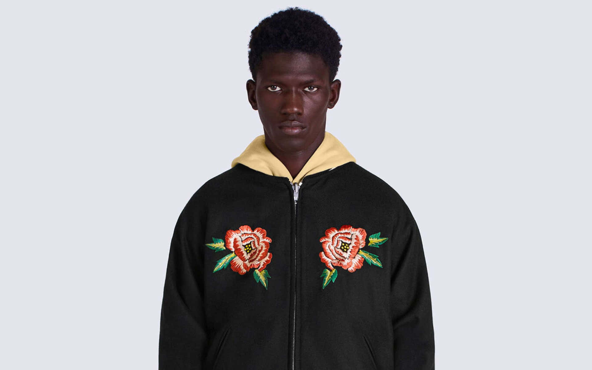 Mens Fashion 桌布 壁紙 — 一名身穿黑色拉鍊連帽上衣、胸前有鮮豔刺繡玫瑰的男子，站在純淺灰色背景前。