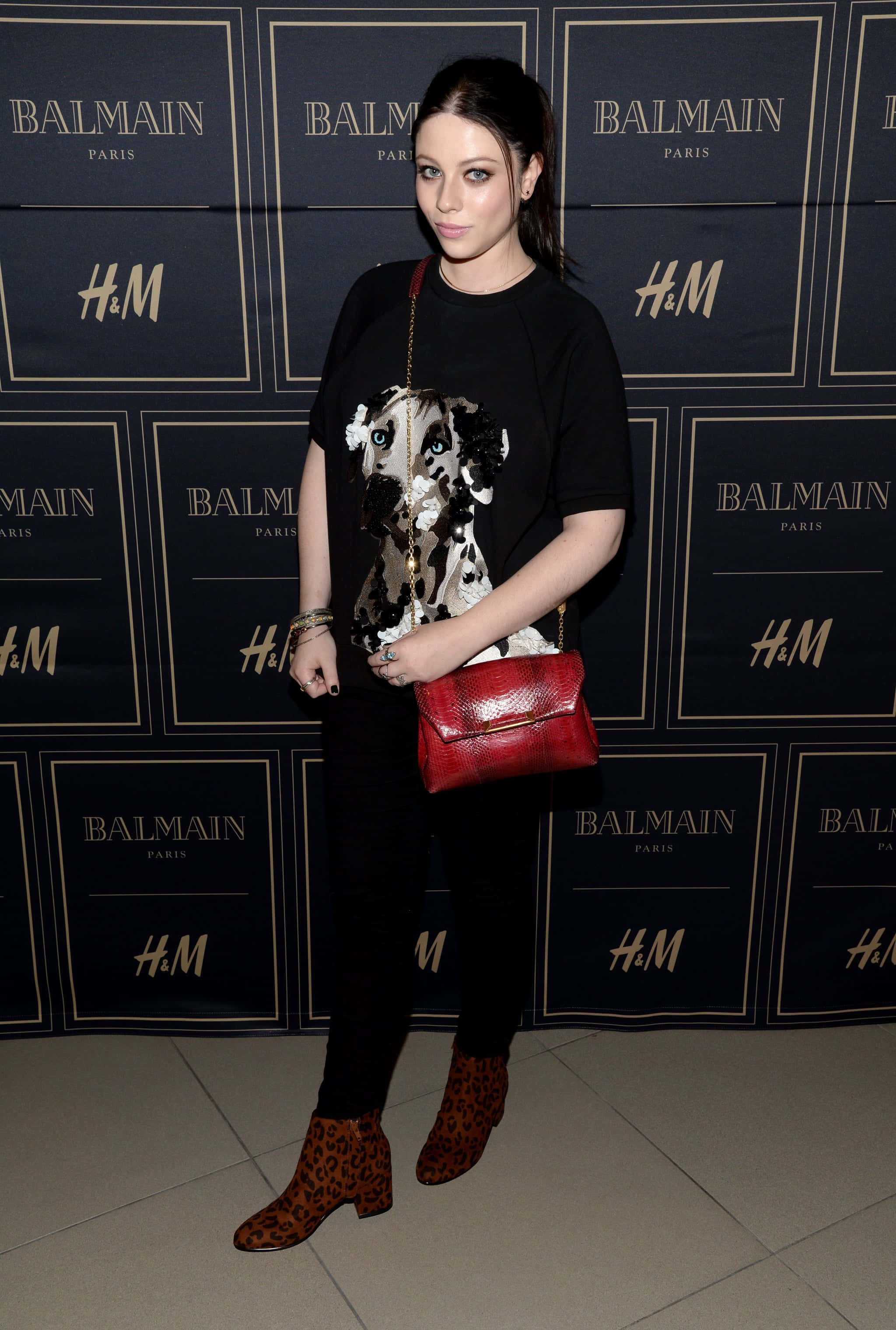 michelle trachtenberg 桌布 壁紙 — 一名身穿黑色服裝、豹紋靴和紅色手提包的女性在活動中站在品牌背景前。