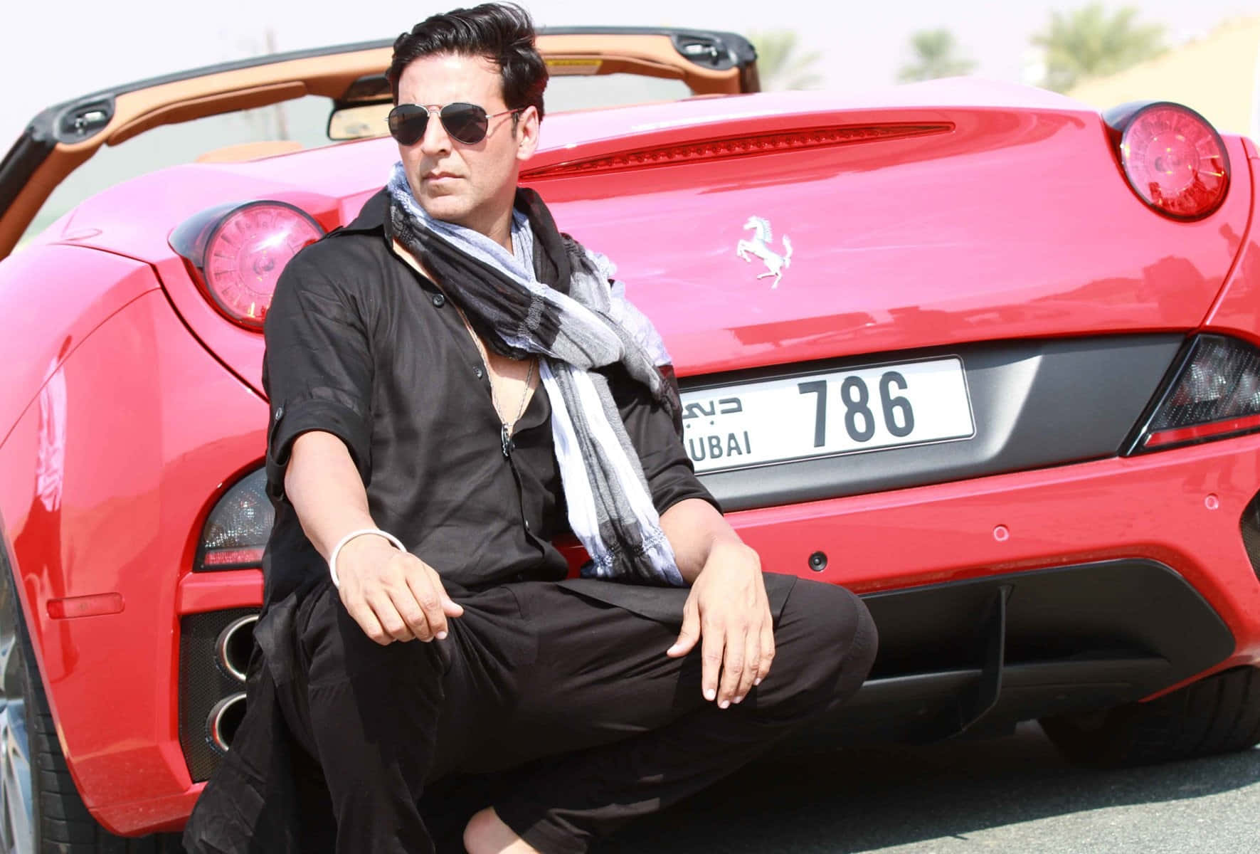 Akshay Kumar 桌布 壁紙 — 一名身穿黑色服裝和太陽眼鏡的男子自信地蹲在掛有杜拜車牌的鮮紅色法拉利敞篷車旁。
