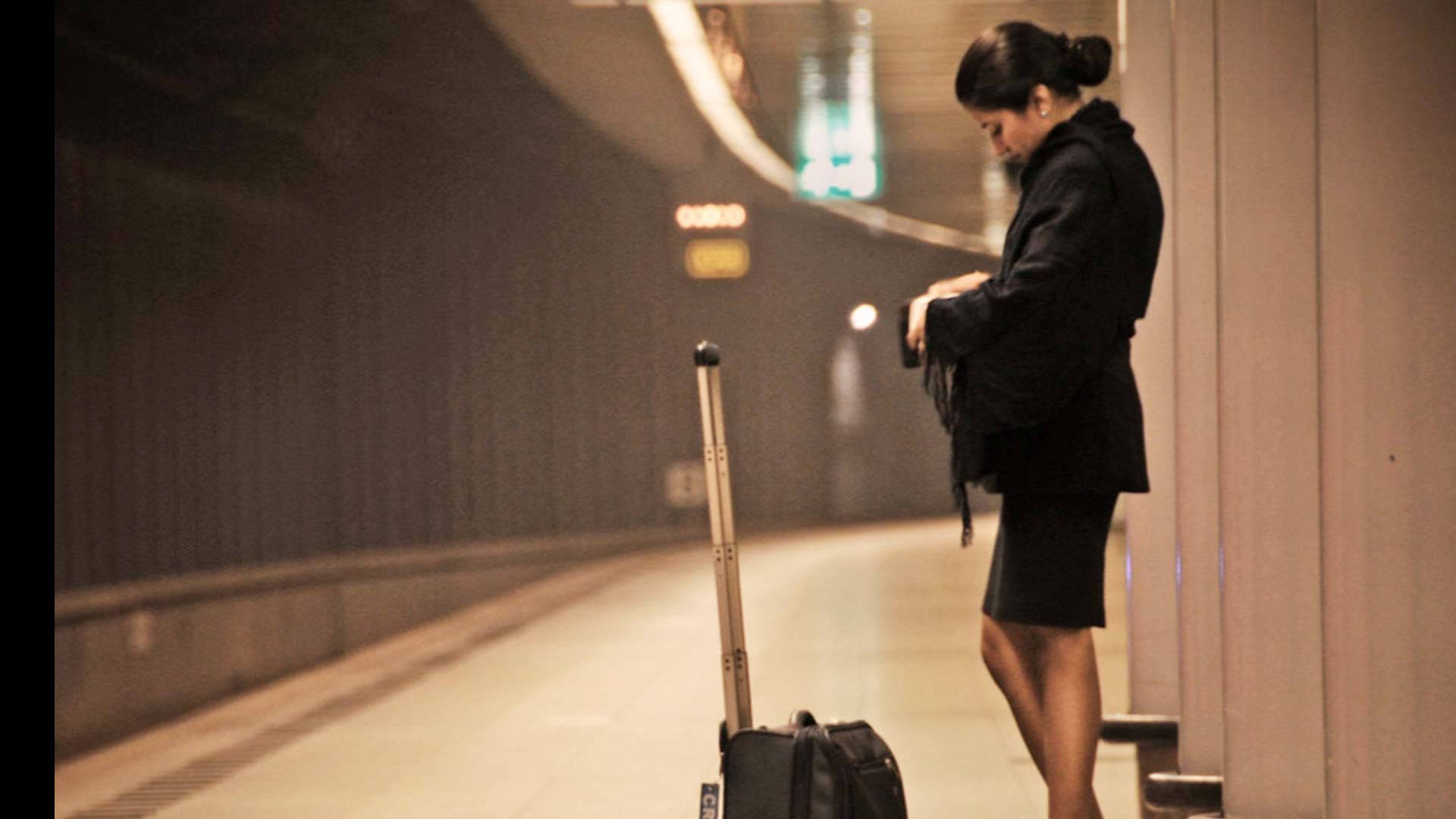 Airport 桌布 壁紙 — 一名身穿黑色服裝的女性獨自站在燈光昏暗的地鐵站月台上，旁邊放著她的滾輪行李箱，低頭看著手機。
