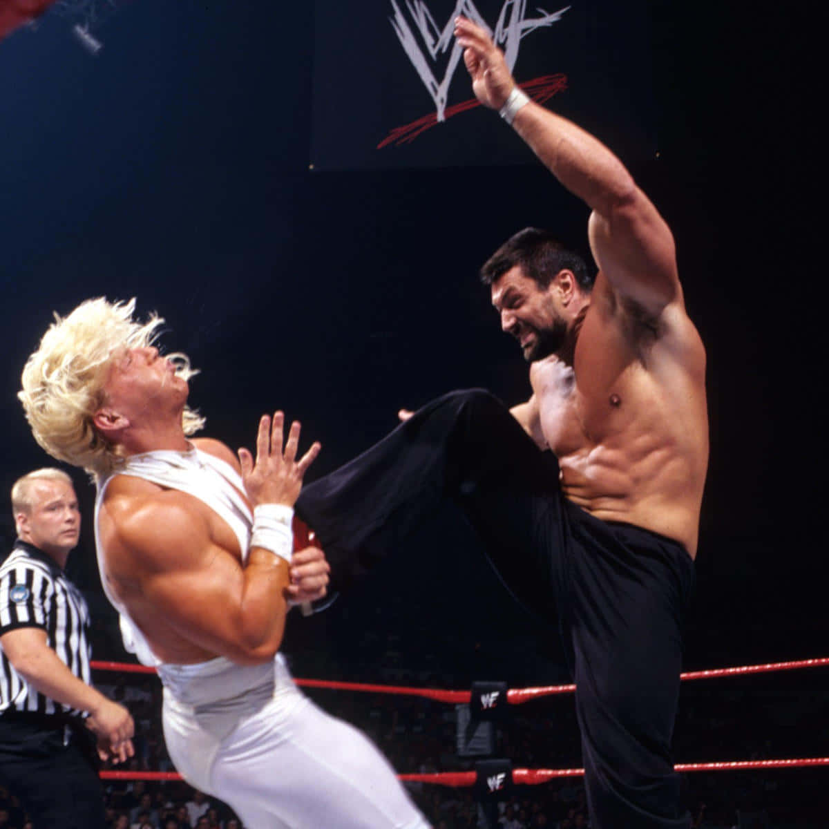 WWE 桌布 壁紙 — 一名身穿黑色服裝的肌肉發達摔角手在明亮的場館燈光下，於WWE比賽中跳起向身穿白色的金髮對手踢出高踢。