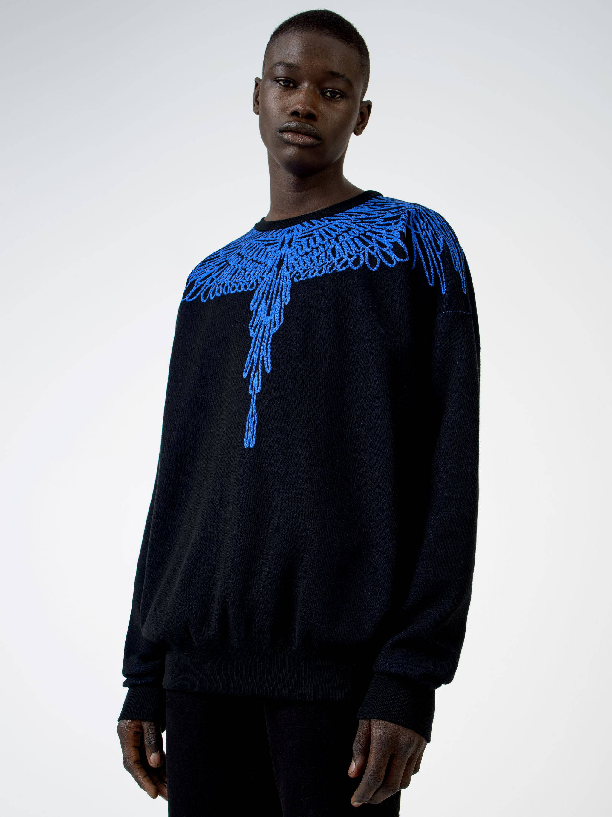 Clothes 桌布 壁紙 — 一名身穿黑色毛衣、肩上帶有醒目藍色刺繡翅膀圖案的男人，站在純白色背景前。