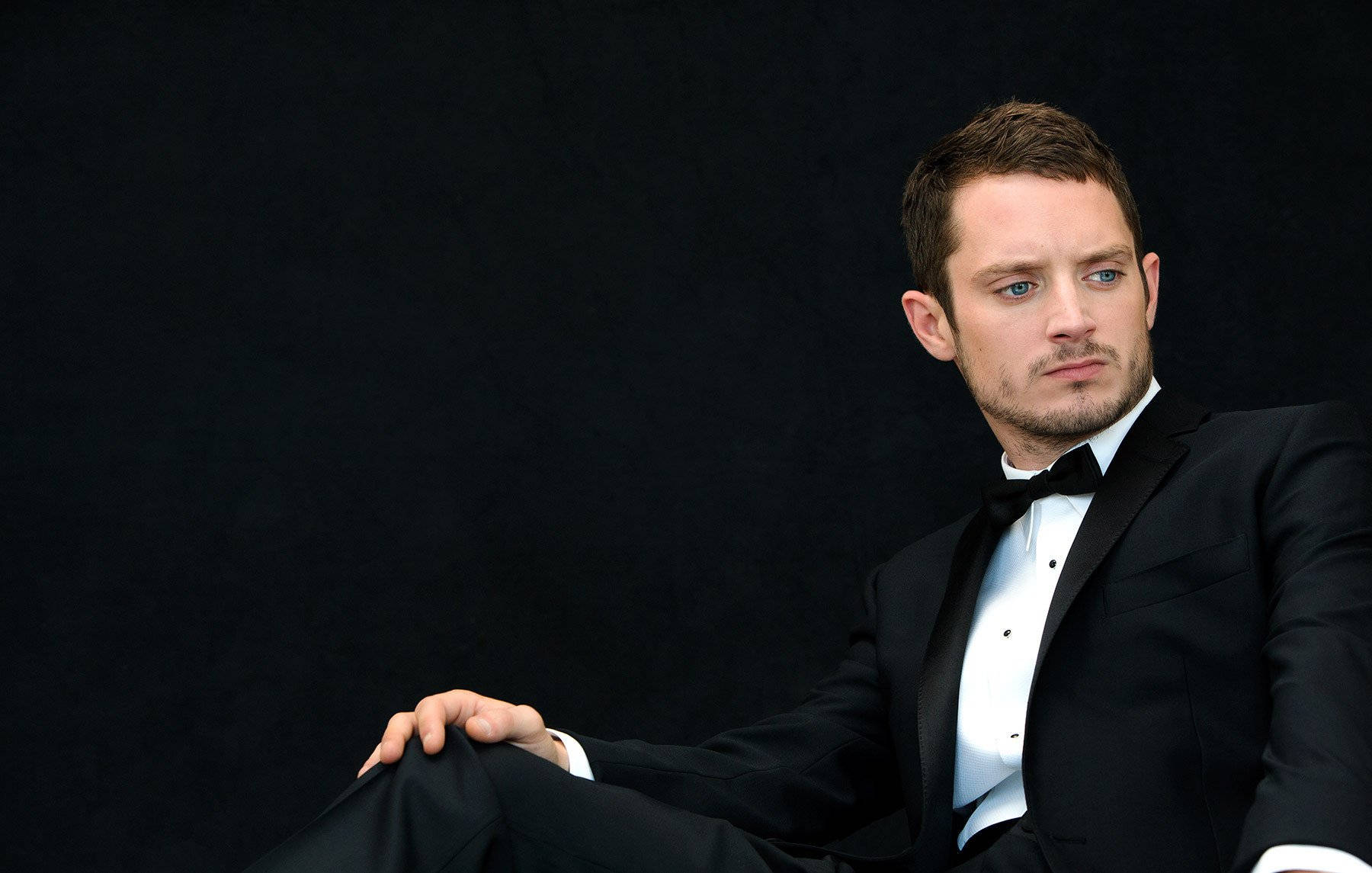 Celebrity 桌布 壁紙 — 一名身穿黑色燕尾服、打著蝴蝶結的男子自信地坐在純黑色背景前，他銳利的藍色眼睛直視觀眾。