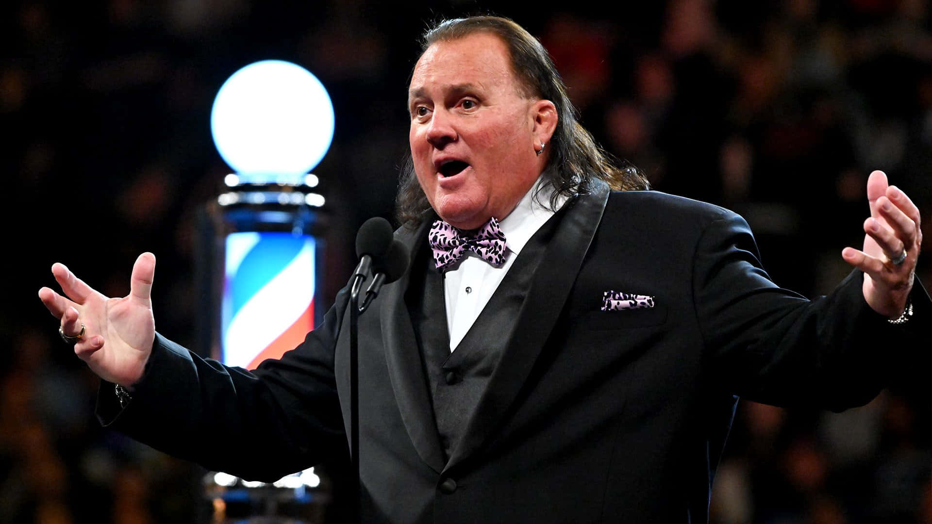 WWE 桌布 壁紙 — 一名身穿黑色燕尾服、繫著圖案領結的男子，在燈光璀璨的舞台上對著麥克風說話，並做出戲劇性的手勢。