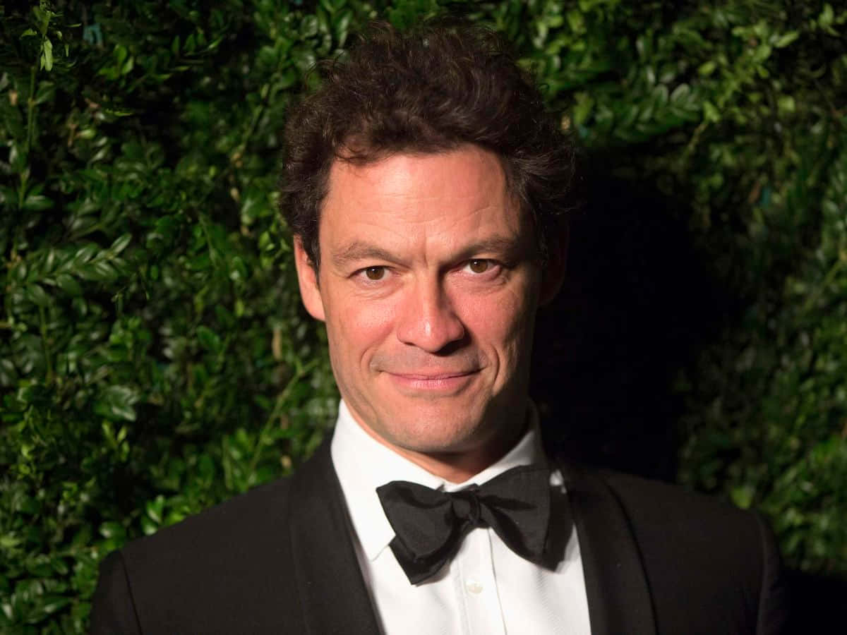 Dominic West 桌布 壁紙 — 一名身穿黑色燕尾服和領結的男子，在茂密的綠色樹籬背景下溫暖地微笑。