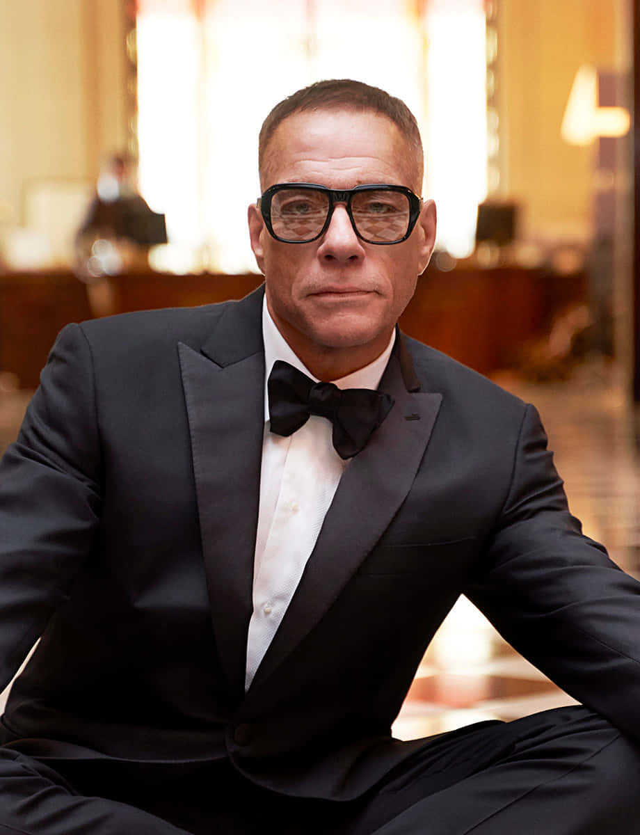 jean claude van damme 桌布 壁紙 — 一名身穿黑色燕尾服和領結的男子，戴著粗黑框眼鏡，自信地坐在溫暖燈光下的優雅室內。