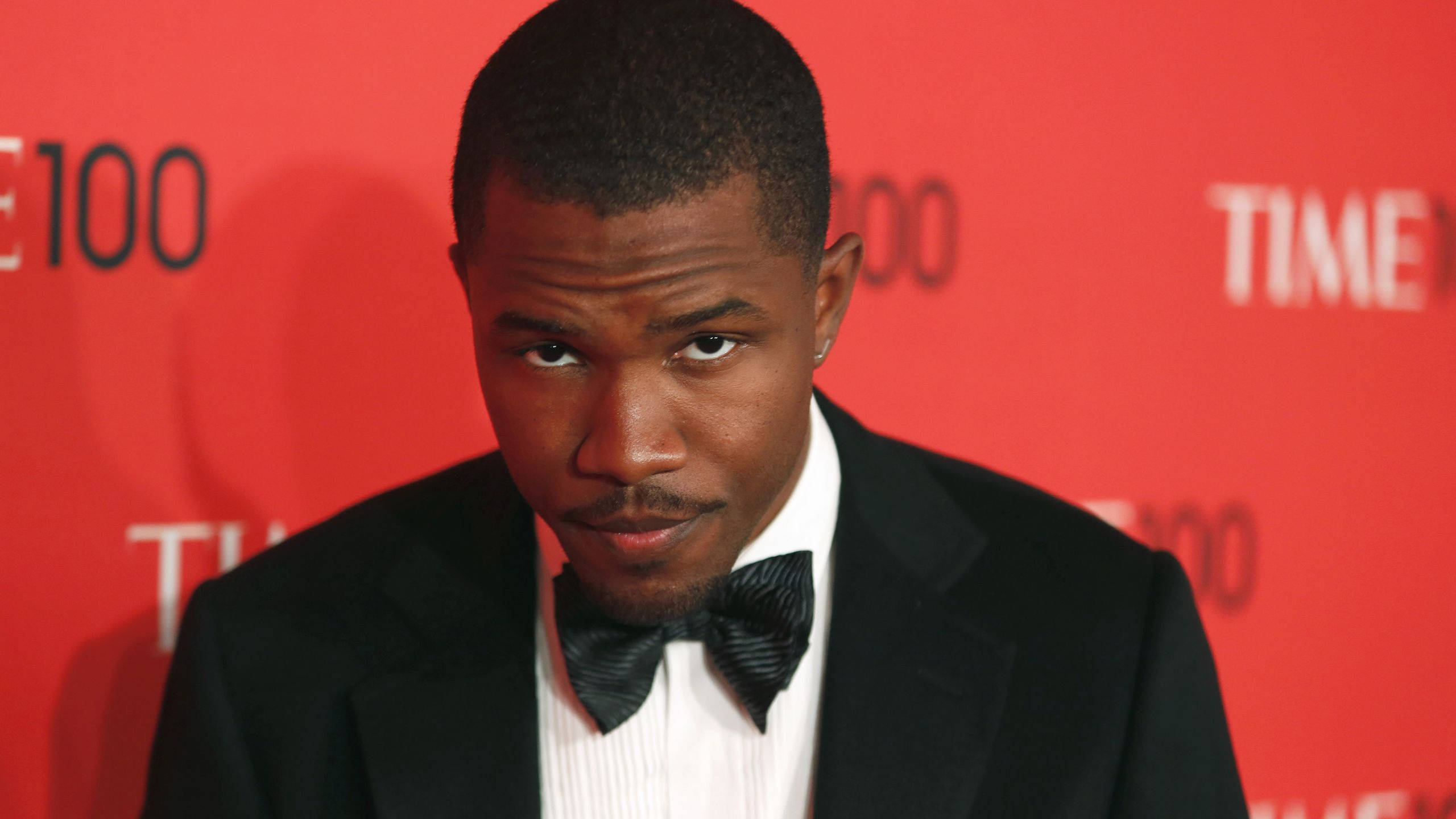 Frank Ocean 桌布 壁紙 — 一名身穿黑色燕尾服和領結的男子在紅色TIME 100活動背景下專注地凝視鏡頭。