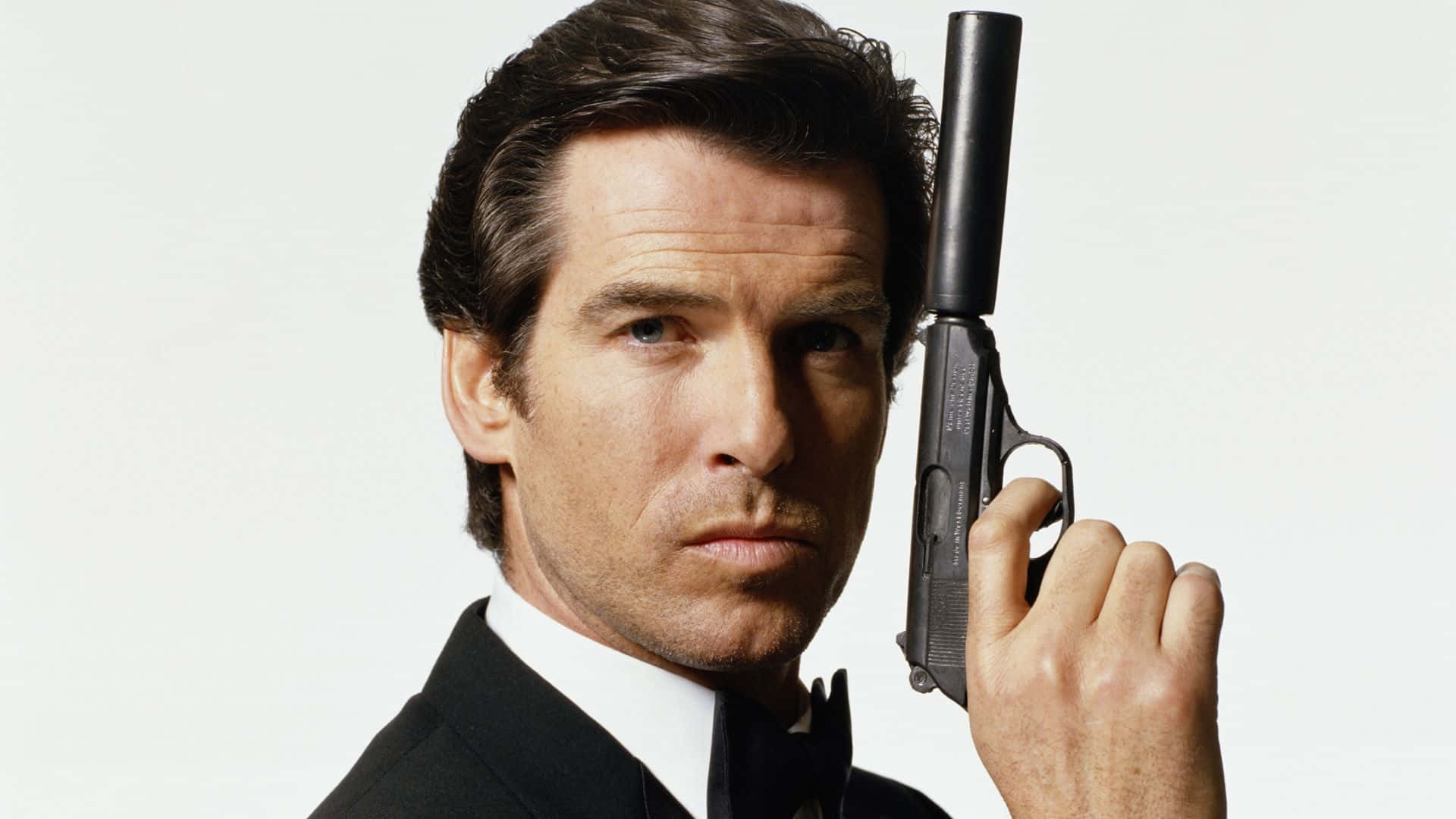 pierce brosnan 桌布 壁紙 — 一名身穿黑色燕尾服的男人，在純白背景前手持消音手槍，表情嚴肅。