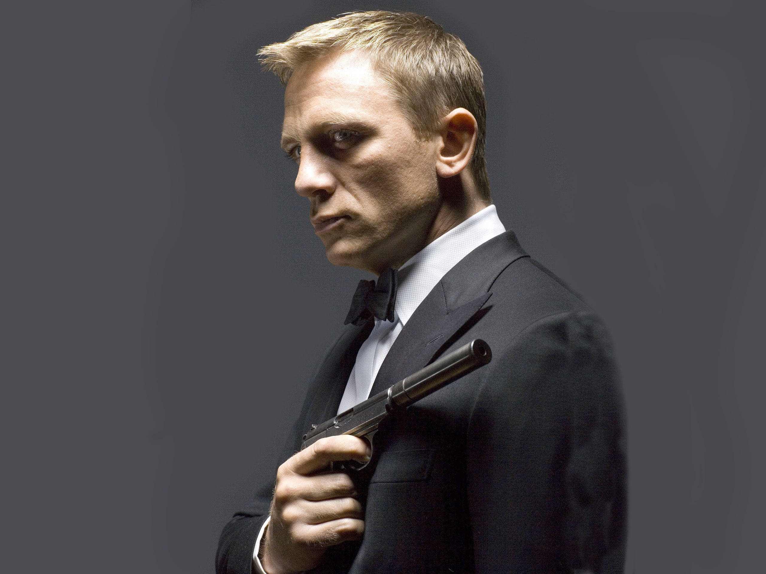 Daniel Craig 桌布 壁紙 — 一名身穿黑色燕尾服的男子，在深灰色背景下持槍，表情嚴肅。