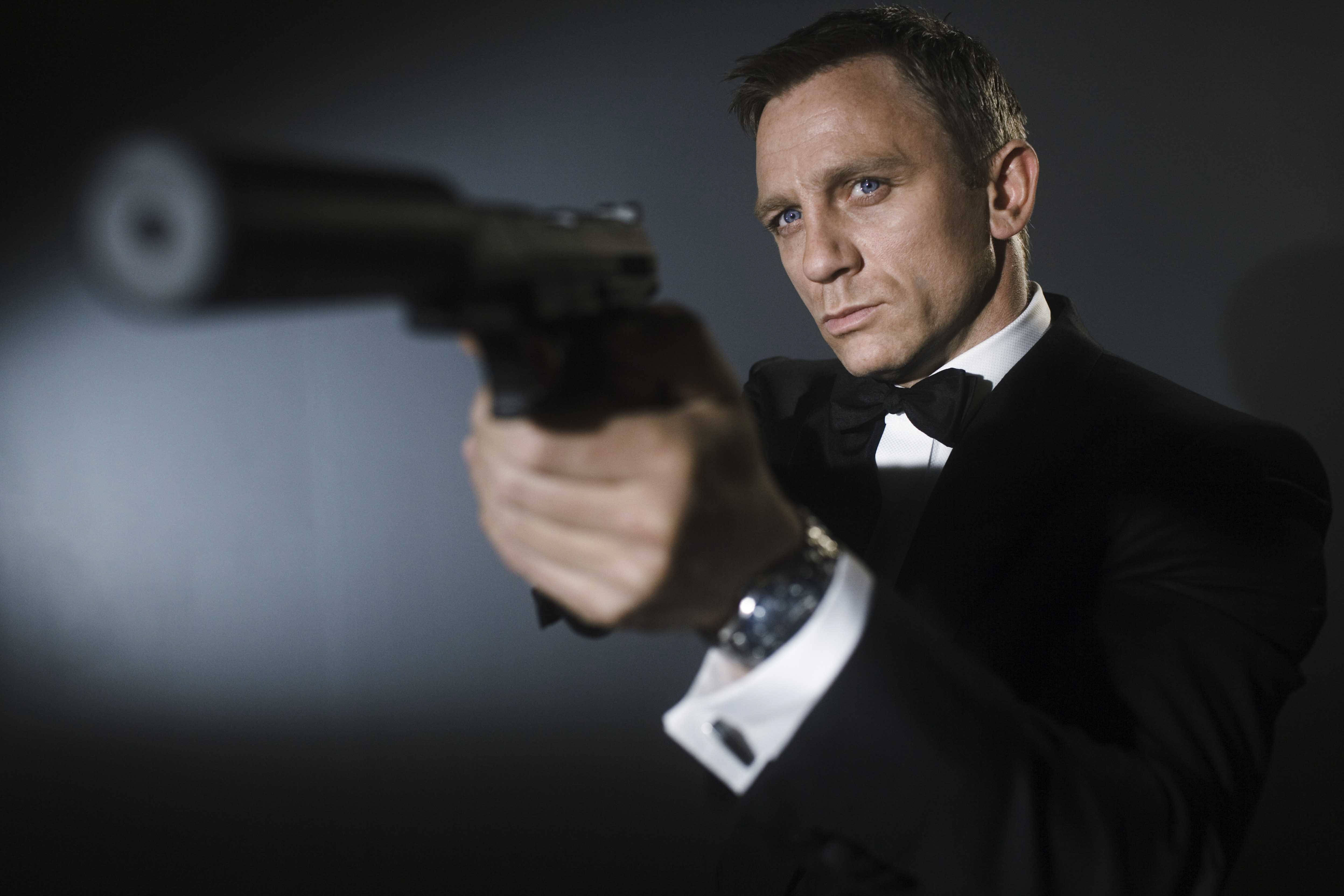 Daniel Craig 桌布 壁紙 — 一名身穿黑色燕尾服的男子在黑暗的攝影棚背景下，專注地瞄準一把消音手槍。