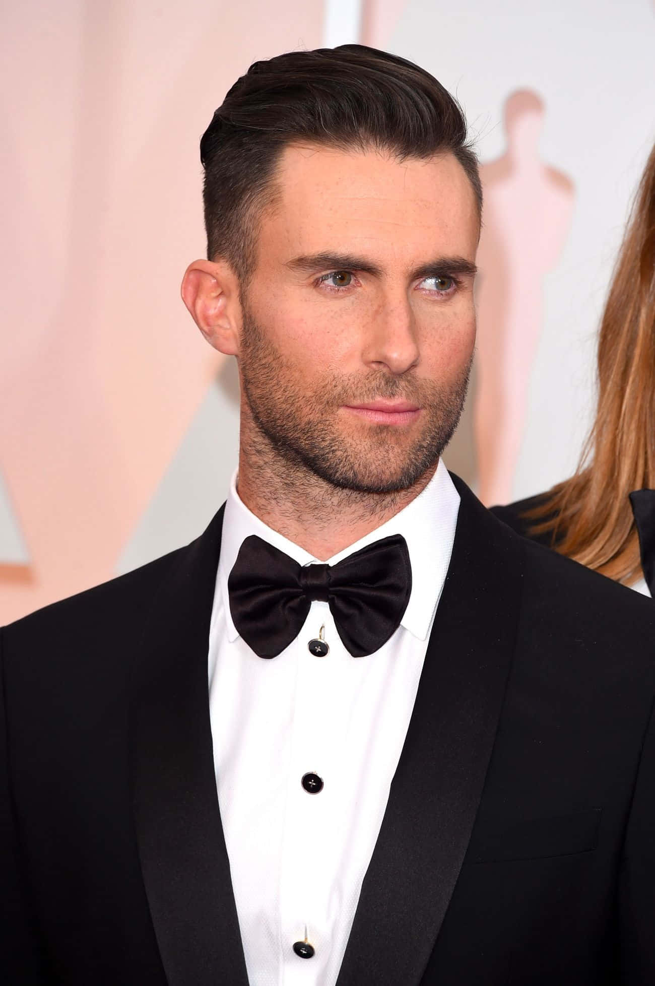 Adam Levine 桌布 壁紙 — 一名身穿黑色燕尾服配蝴蝶結的男子，在柔和的粉紅色背景的紅地毯活動中自信地站立。