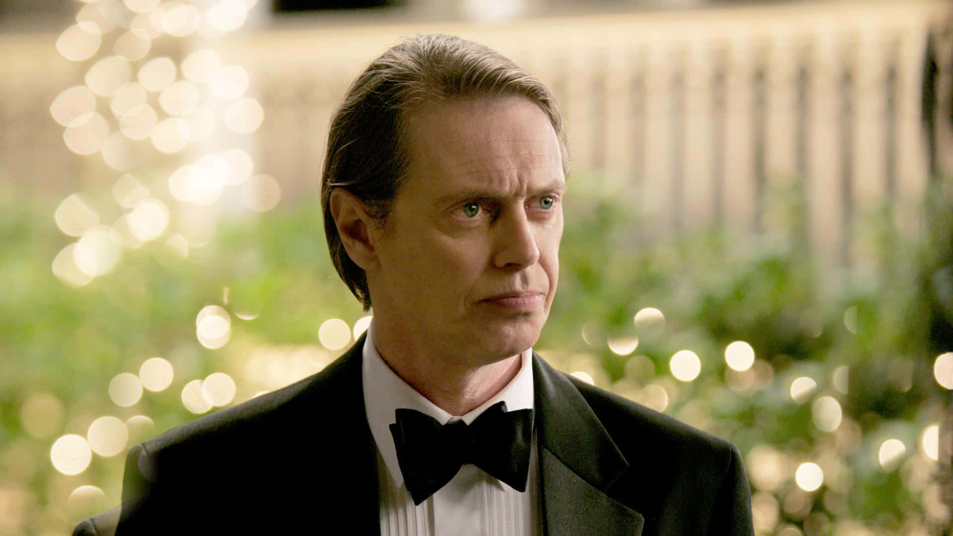 steve buscemi 桌布 壁紙 — 一名身穿黑色燕尾服配蝴蝶結的男子，在模糊的金色燈光和綠植背景下若有所思地凝視。