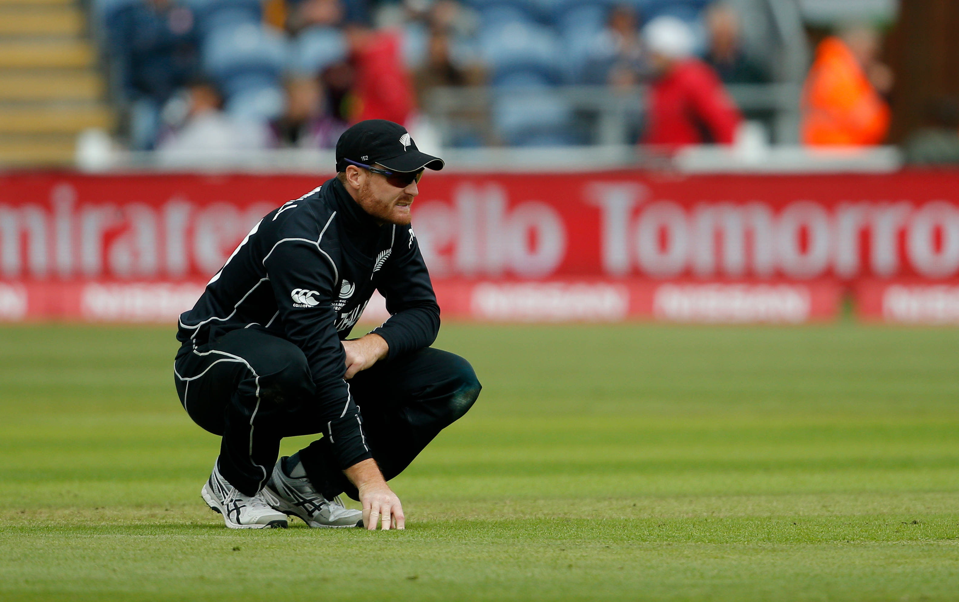 martin guptill 桌布 壁紙 — 一名身穿黑色球衣的板球運動員在充滿活力的綠色場地上低蹲，背景是模糊的體育場座位。