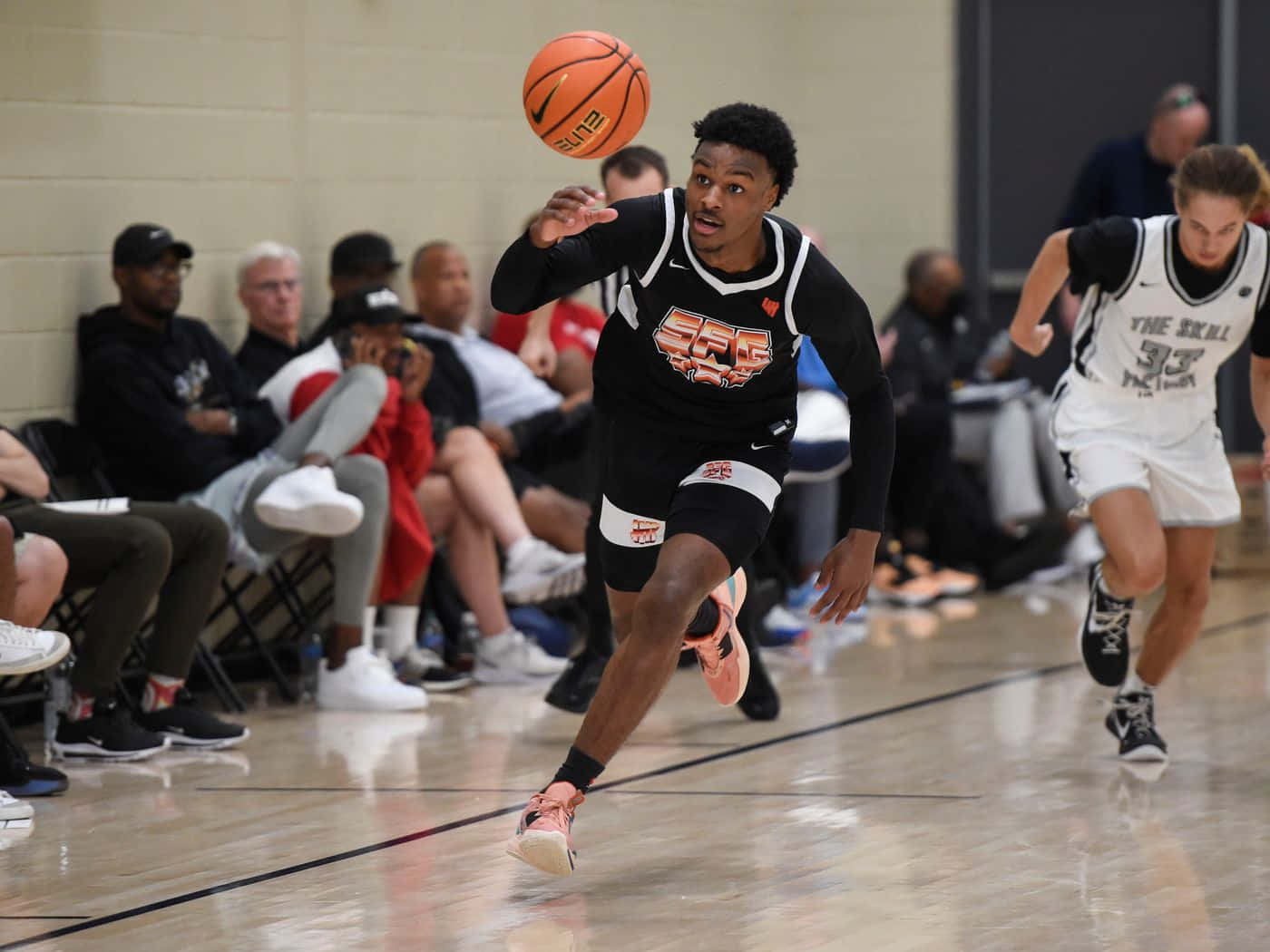 Bronny James 桌布 壁紙 — 一名身穿黑色球衣的籃球運動員在室內比賽中運球，同時被一名身穿白色球衣的防守球員追趕。