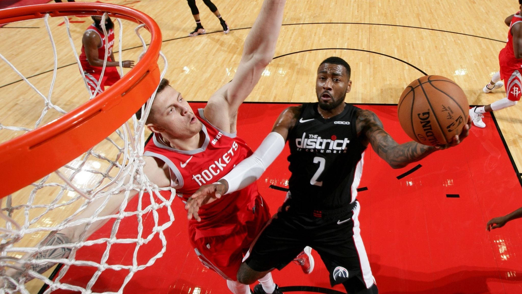 John Wall 桌布 壁紙 — 一名身穿黑色球衣的籃球運動員在明亮的場館燈光下，向籃框推進，同時被一名紅色球衣的球員防守。