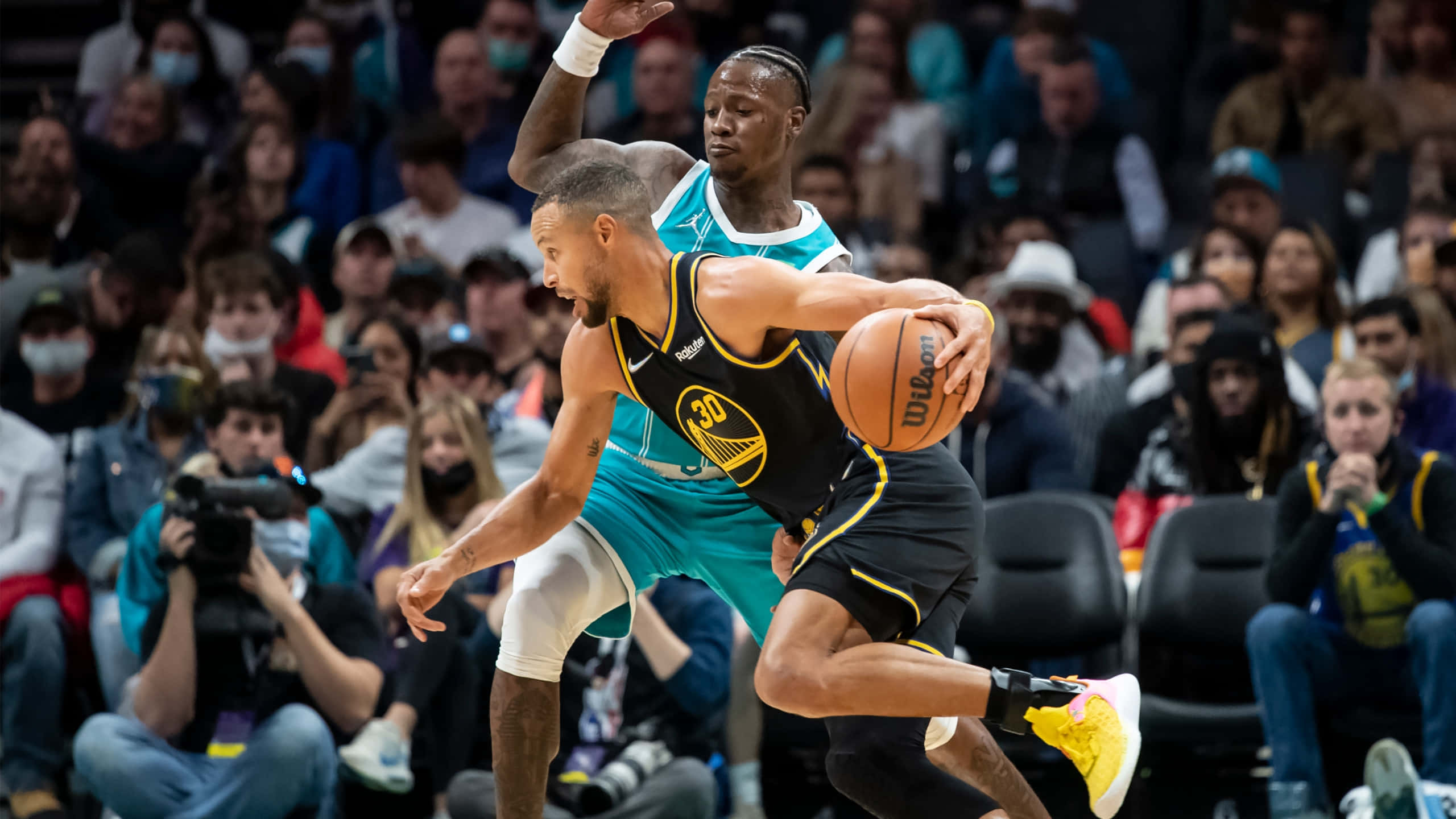 NBA 桌布 壁紙 — 一名身穿黑色球衣的籃球運動員在激烈的比賽中運球越過一名青綠色防守球員，背景是座無虛席的觀眾。