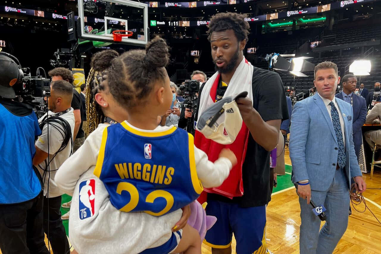 Andrew Wiggins 桌布 壁紙 — 一名身穿黑色球衣的籃球運動員在燈光明亮的球場上與一名身穿藍色Wiggins 22球衣的小女孩熱情互動。