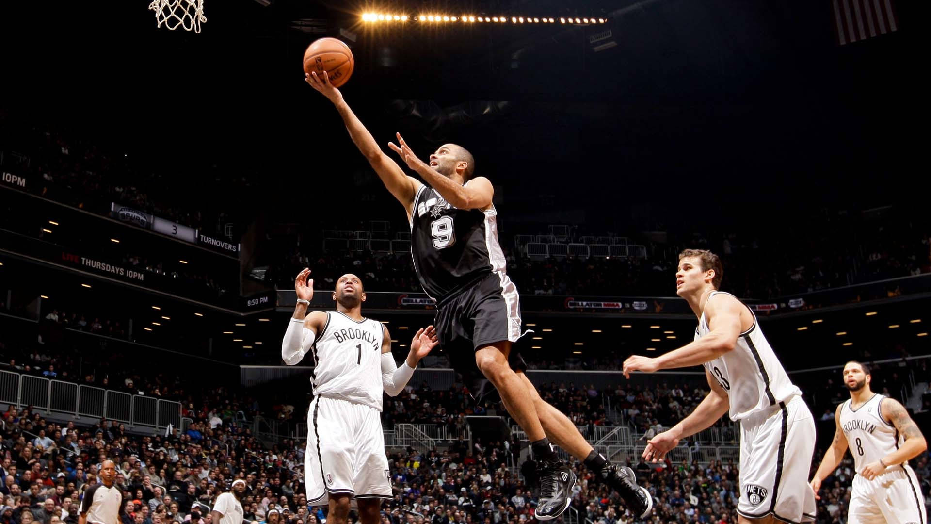 Tony Parker 桌布 壁紙 — 一名身穿黑色球衣的籃球運動員高高躍起準備灌籃，身穿白色球衣的防守球員在明亮的場館燈光下試圖封阻他。