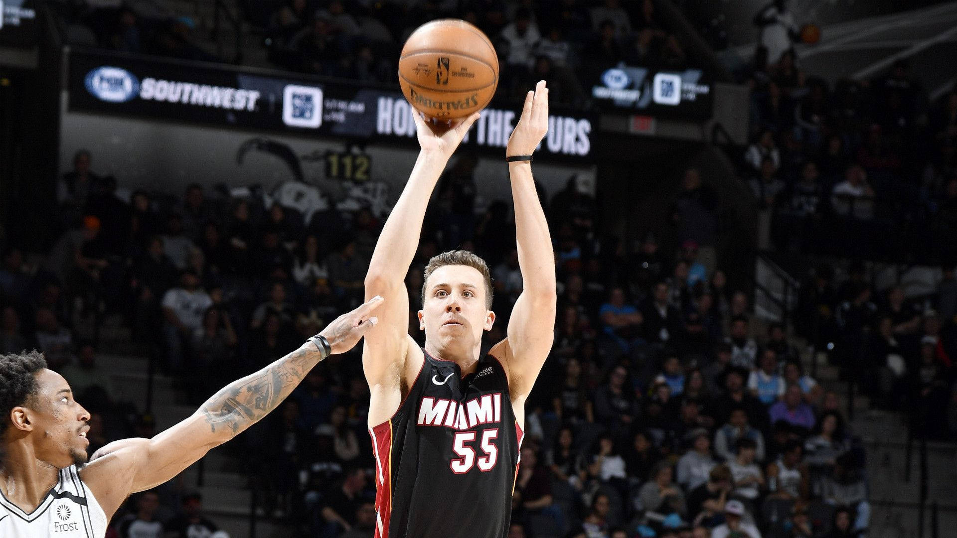 duncan robinson 桌布 壁紙 — 一名身穿黑色球衣的邁阿密熱火球員正在投籃，而一名身穿白色球衣的防守球員在擠滿觀眾的場館中試圖封阻他。