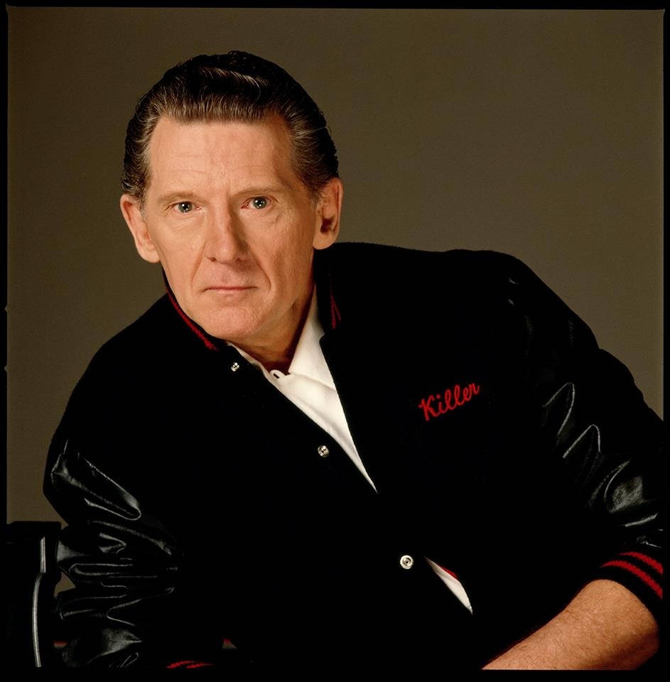 Jerry Lee Lewis 桌布 壁紙 — 一名身穿黑色皮夾克、紅色飾邊、紅色刺繡「Killer」字樣的男人，坐在柔和的棕色背景前。