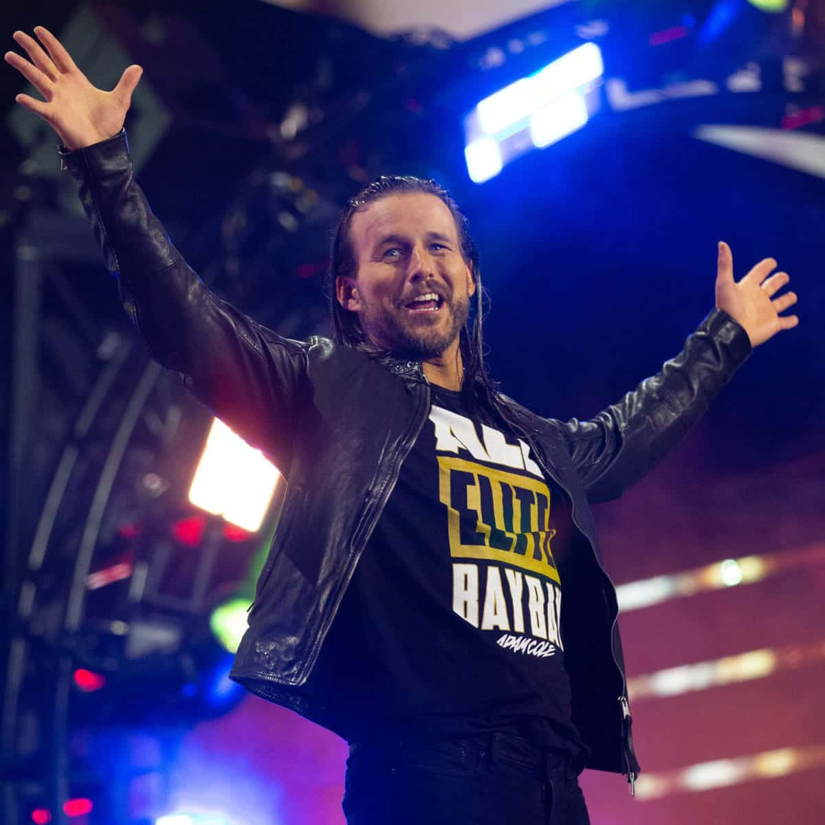 professional 桌布 壁紙 — 一名身穿黑色皮夾克和「All Elite Wrestling」T恤的摔角手，在明亮的舞台燈光下高舉雙臂慶祝勝利。