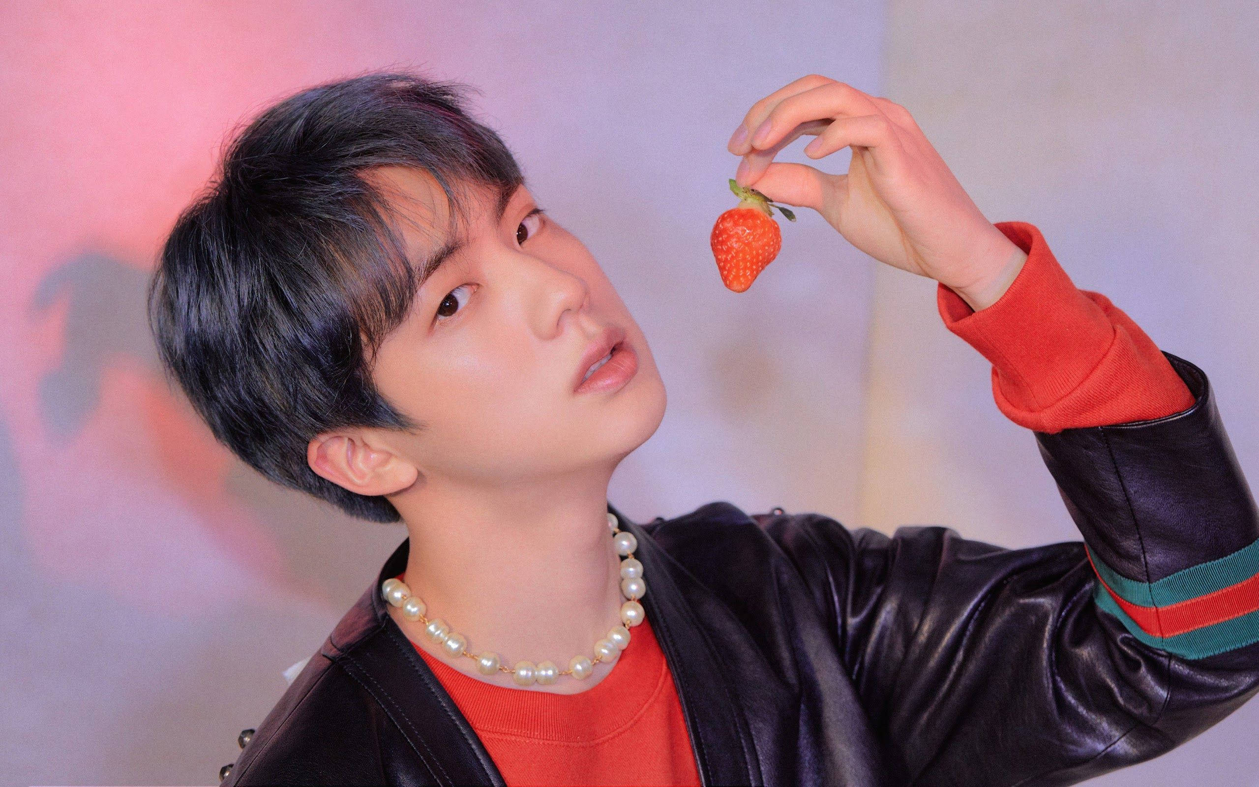 kim seok jin 桌布 壁紙 — 一名身穿黑色皮夾克和珍珠項鍊的年輕男子，在柔和的粉紅和紫色漸變背景前，將草莓靠近臉部。