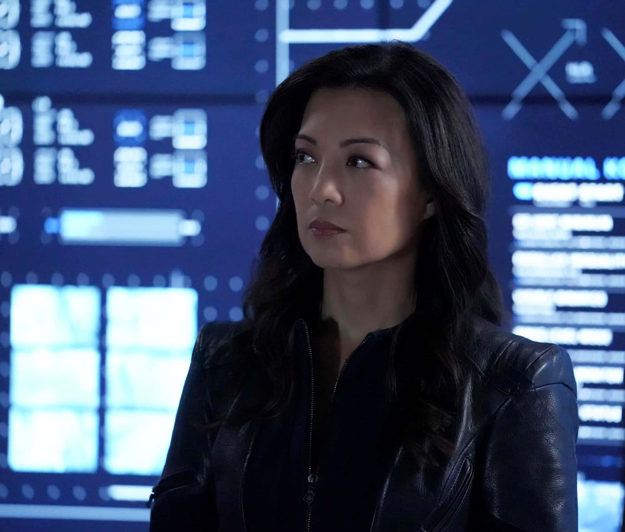 Ming Na Wen 桌布 壁紙 — 一名身穿黑色皮夾克的女子站在高科技控制室中，面前是顯示複雜數據的藍色發光數位螢幕。