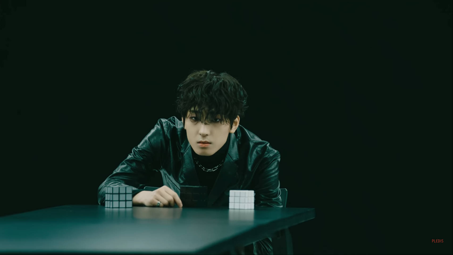 Wonwoo 桌布 壁紙 — 一名身穿黑色皮夾克的年輕男子，在陰暗的綠色燈光下，俯身於一張放有兩個魔術方塊的深色桌子上。