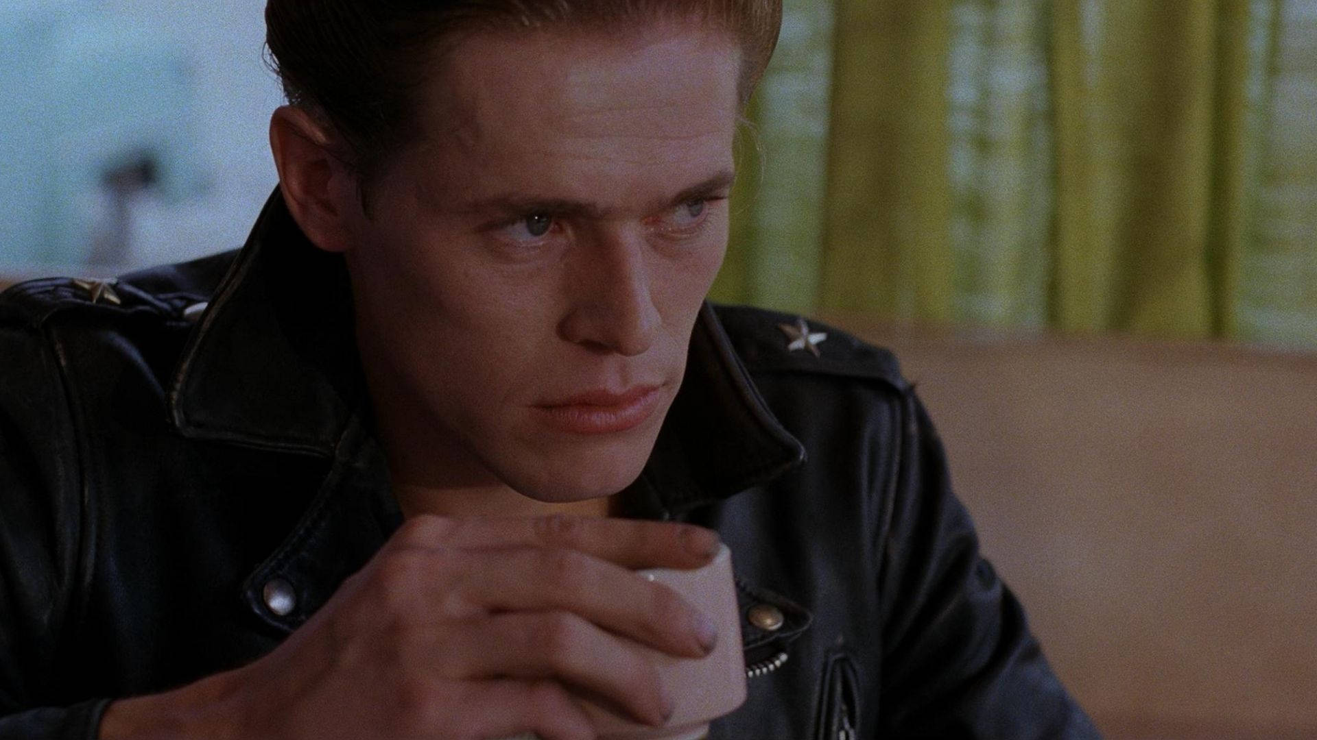Willem Dafoe 桌布 壁紙 — 一名身穿黑色皮夾克的男子，在室內靠近綠色窗簾的地方，手持杯子，表情嚴肅。