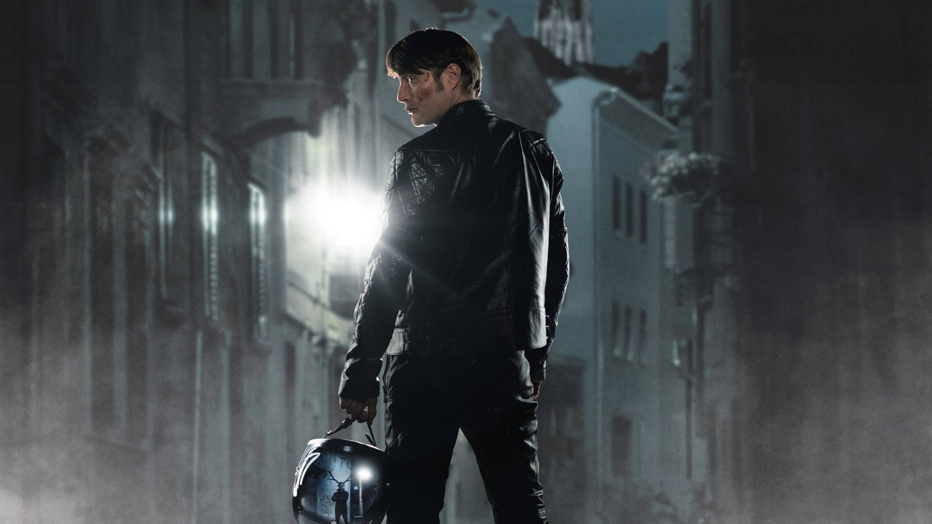 Mads Mikkelsen 桌布 壁紙 — 一名身穿黑色皮夾克的男子在夜晚的霧氣瀰漫的城市街道上行走，在明亮的街燈下拿著頭盔。