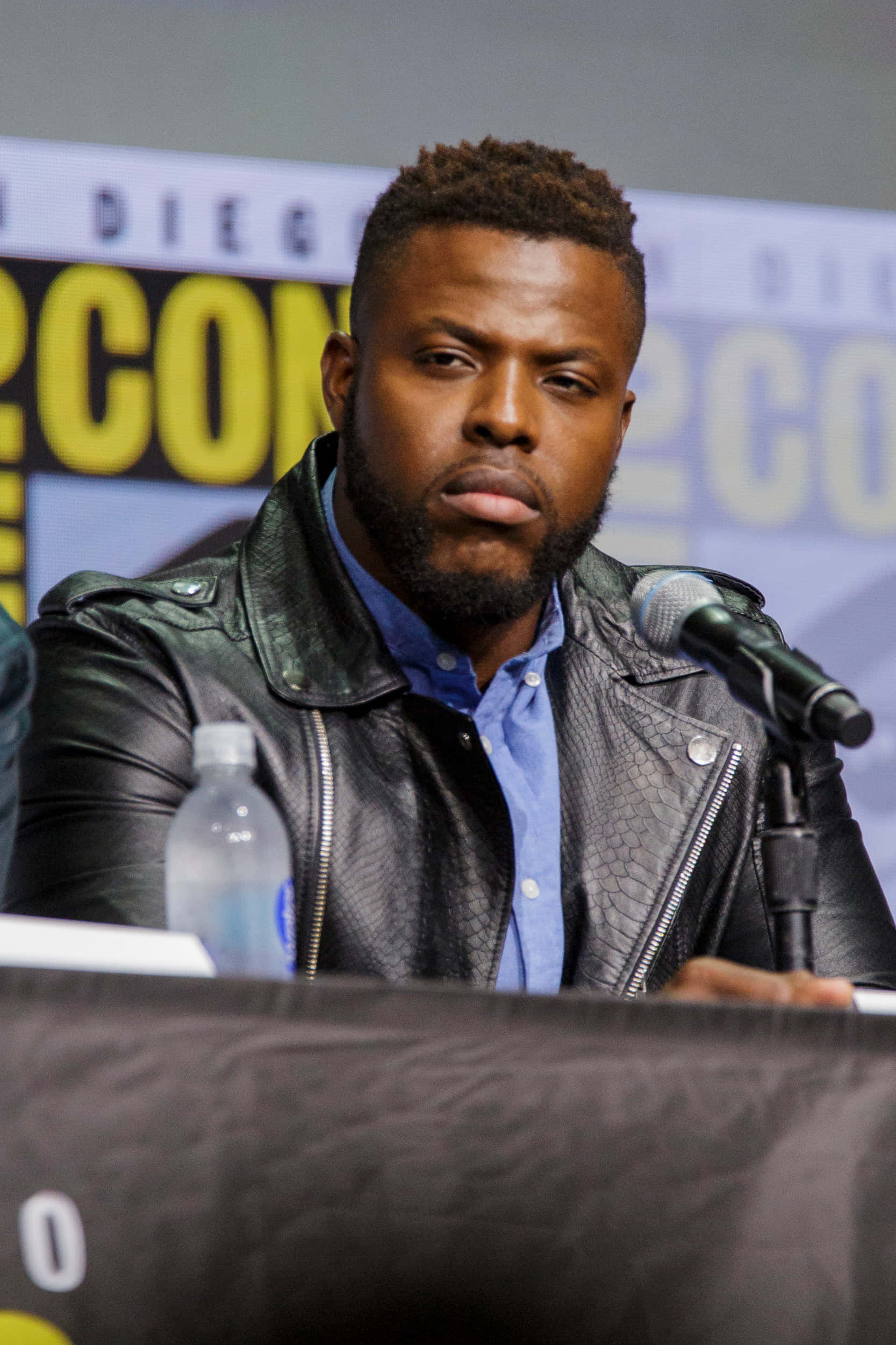 Winston Duke 桌布 壁紙 — 一名身穿黑色皮夾克的男子坐在Comic-Con的座談會桌上，桌上有一個麥克風和一個水瓶。