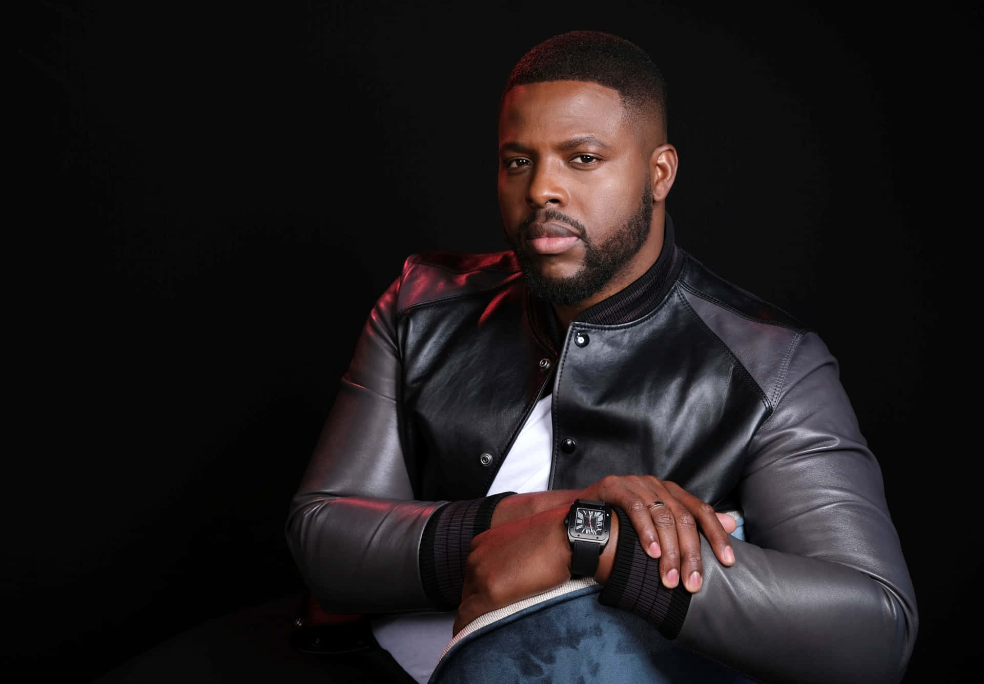Winston Duke 桌布 壁紙 — 一名身穿黑色皮夾克的男子自信地坐在深色背景前，肩上帶有紅色燈光點綴。
