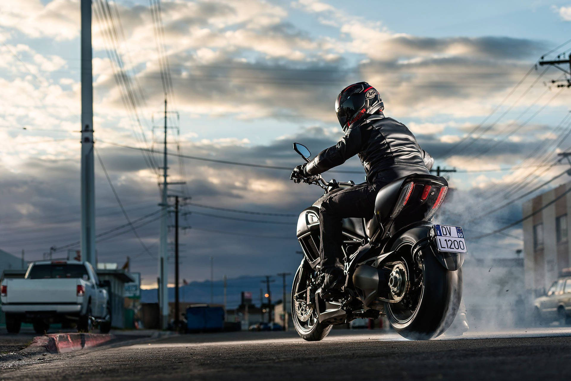 motorbike 桌布 壁紙 — 一名身穿黑色皮衣和頭盔的機車騎士，在黃昏時分騎著一輛時尚的黑色機車行駛在街上，後輪冒出煙霧。