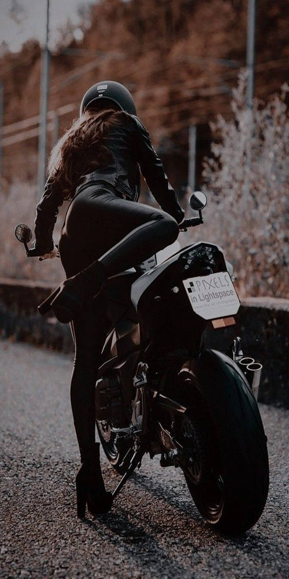  壁紙 — 一名身穿黑色皮革和頭盔的女性，在碎石路上跨坐著一輛時尚摩托車，背景是秋樹。