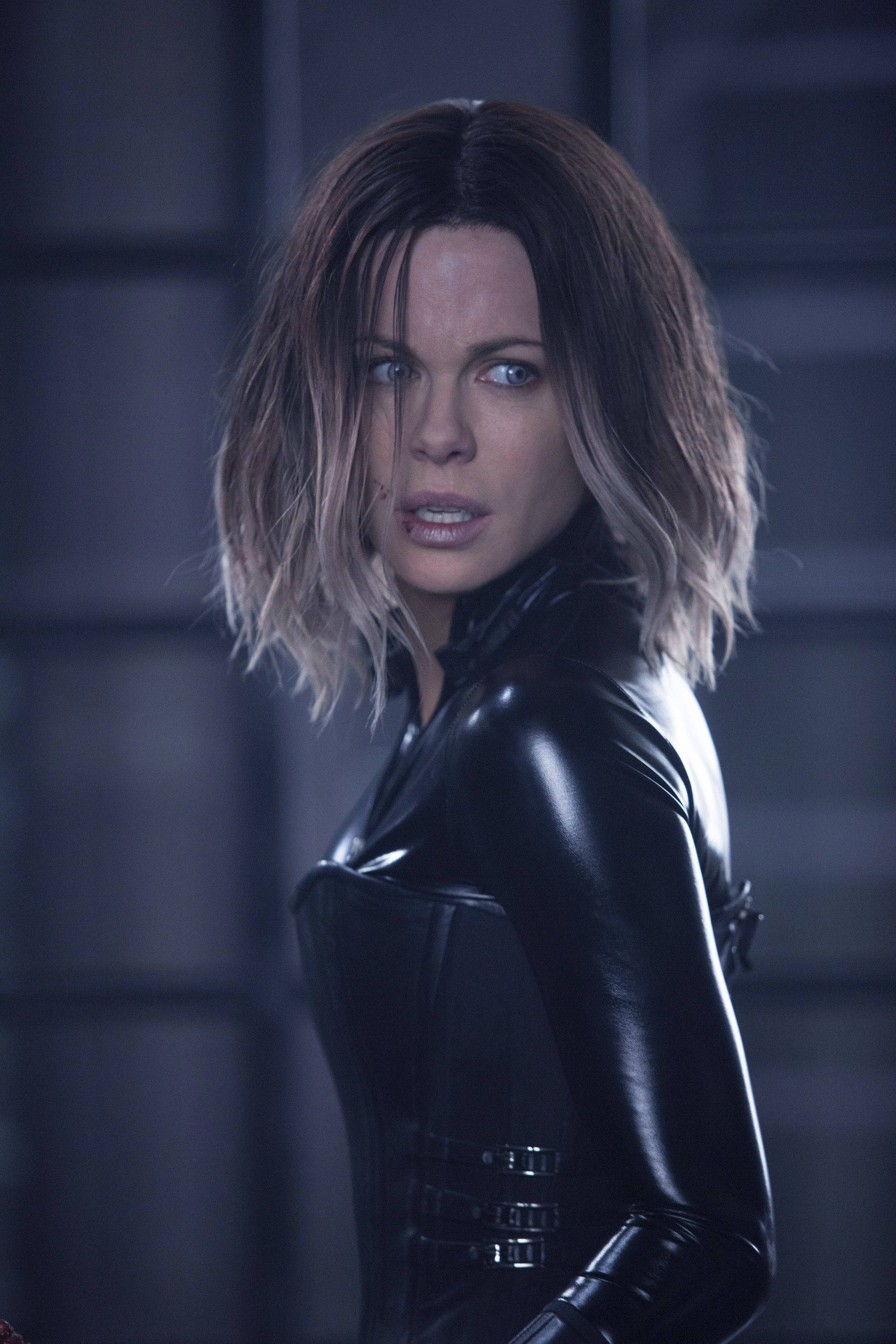 Kate Beckinsale Underworld 桌布 壁紙 — 一名身穿黑色皮革緊身衣的女子，在深灰色瓷磚牆前轉過身來，露出強烈的藍色眼睛。