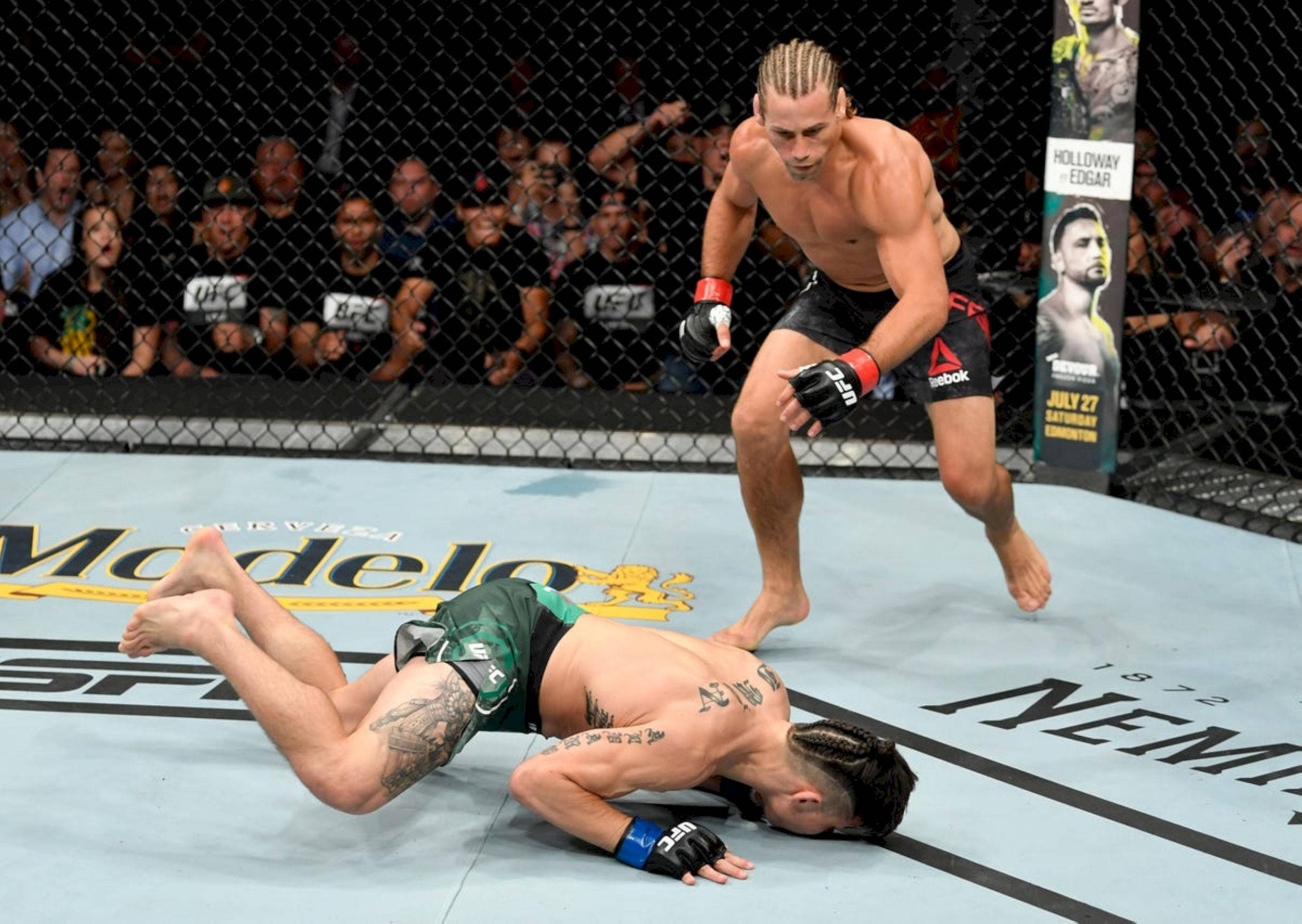 MMA 桌布 壁紙 — 一名身穿黑色短褲的拳手在UFC八角籠內的軟墊上，擊敗對手並站立其上，鐵籠外有歡呼的人群。