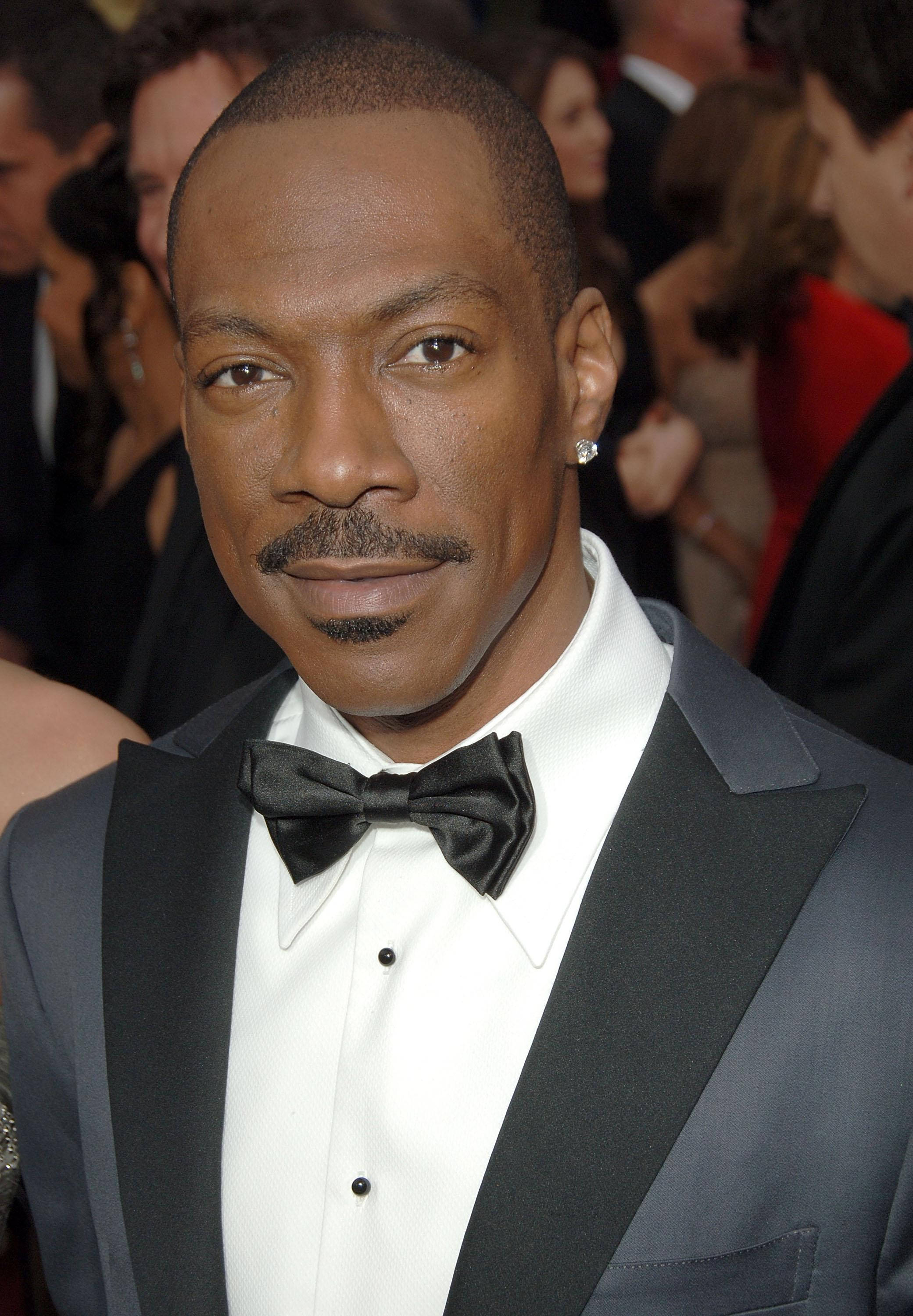 eddie murphy 桌布 壁紙 — 一名身穿黑色禮服和領結的男士特寫肖像，身後有模糊的賓客，在一個華麗的活動中。