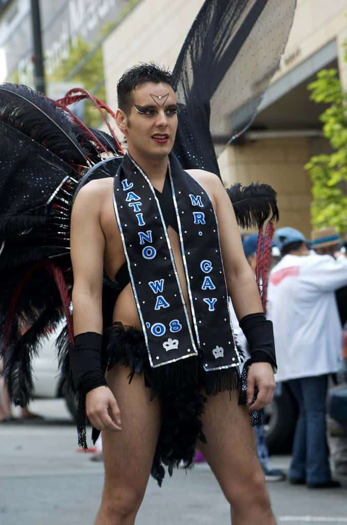 驕傲 桌布 壁紙 — 一名身穿黑色羽毛服裝、佩戴「Mr. Gay Latino 2008」綬帶的男子，在遊行期間自信地在城市街道上擺姿勢。